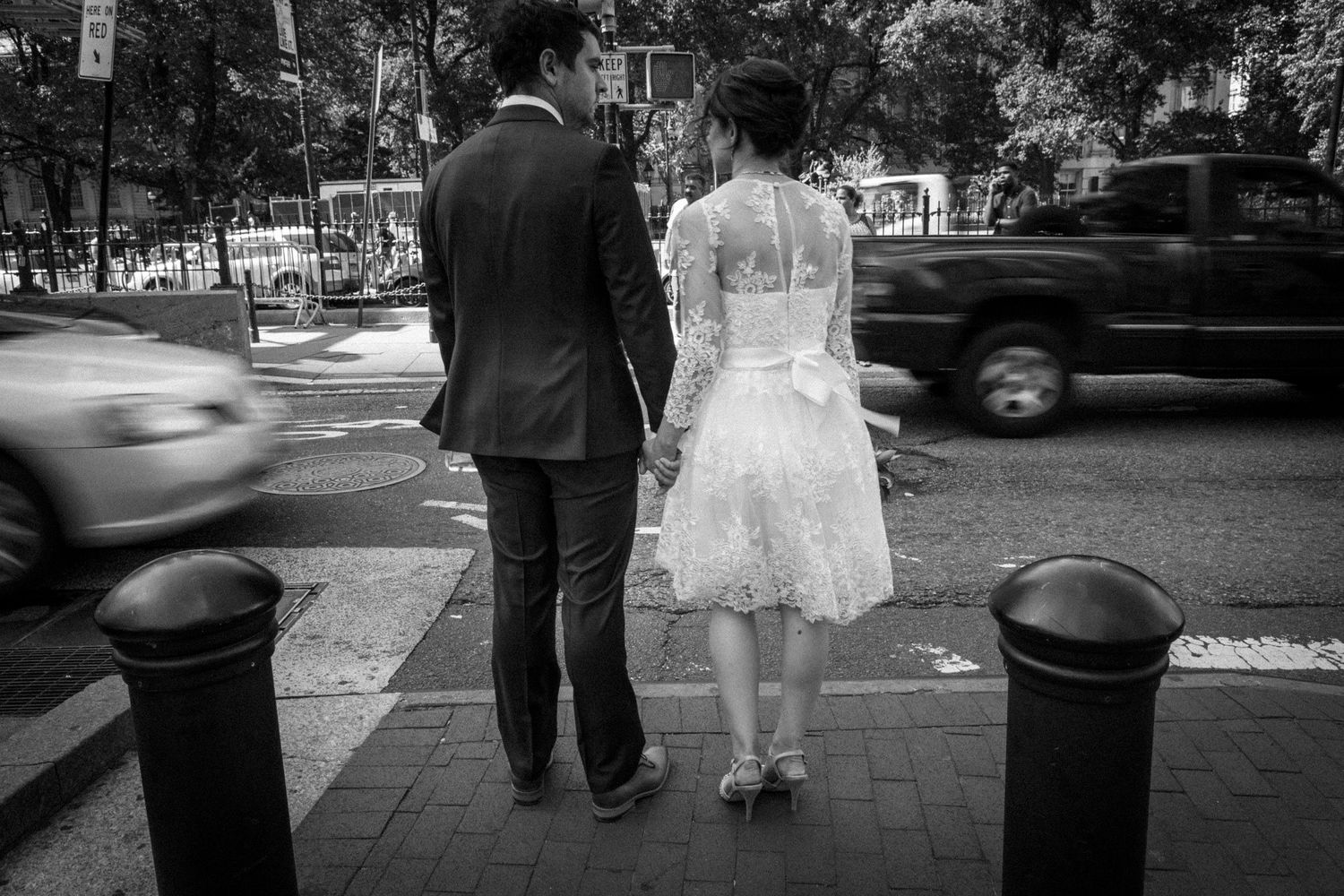 Black and white photo of a bride and groom holding hands on a city sidewalk with traffic passing by.