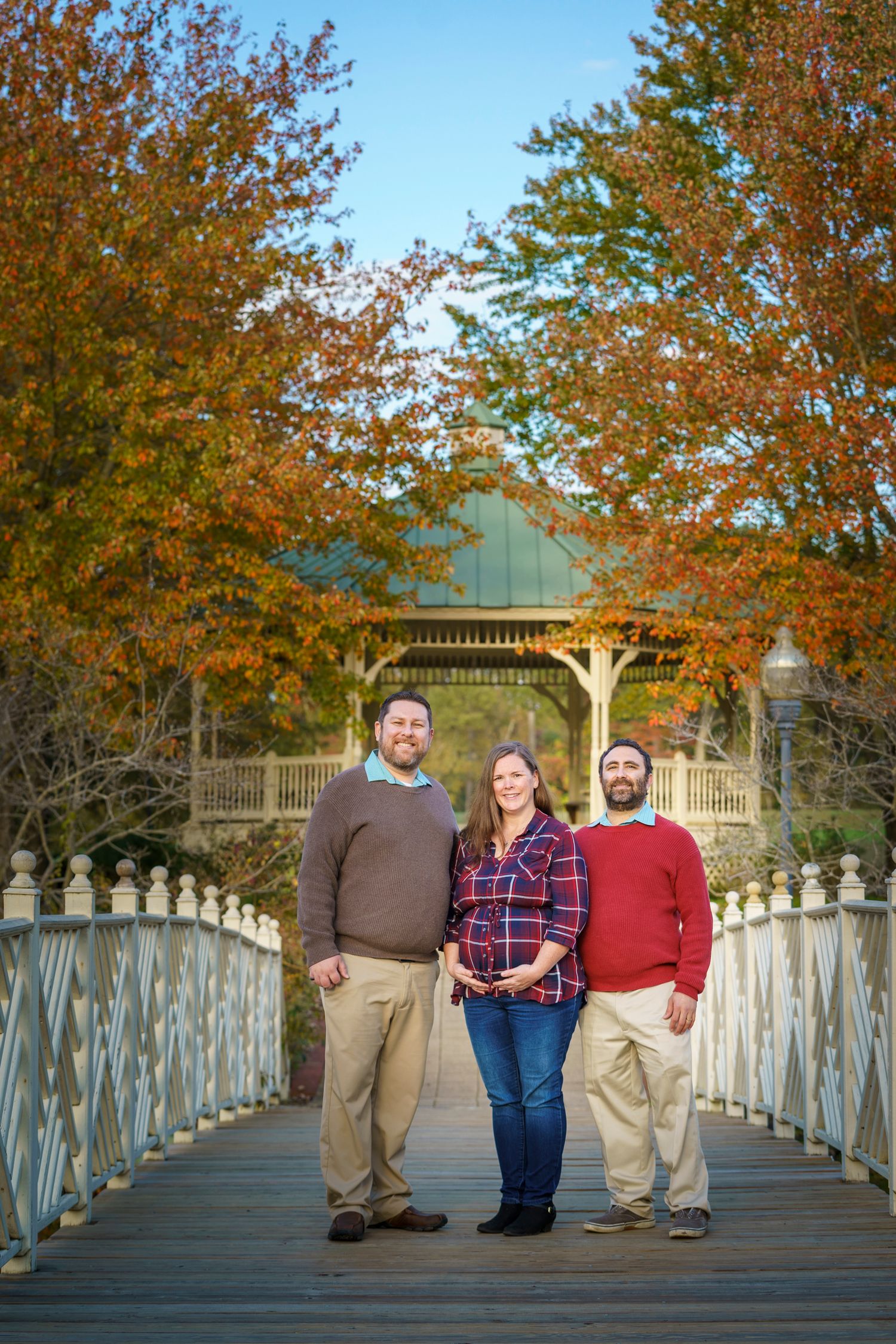 Three people pose on a wooden bridge surrounded by autumn foliage near a gazebo.