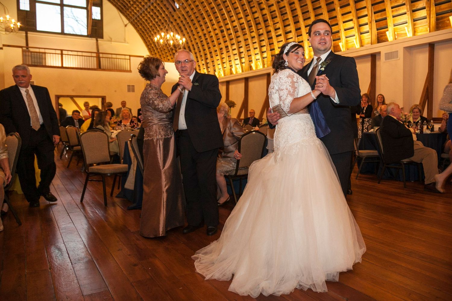 Bride and groom share first dance alongside parents in elegant wood-beamed wedding venue.