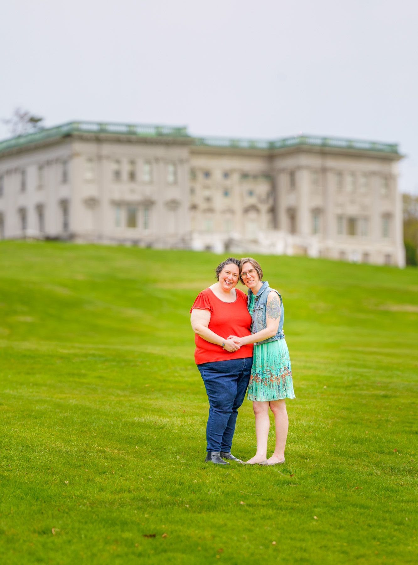Two women embrace on green lawn before grand stone mansion under overcast sky.