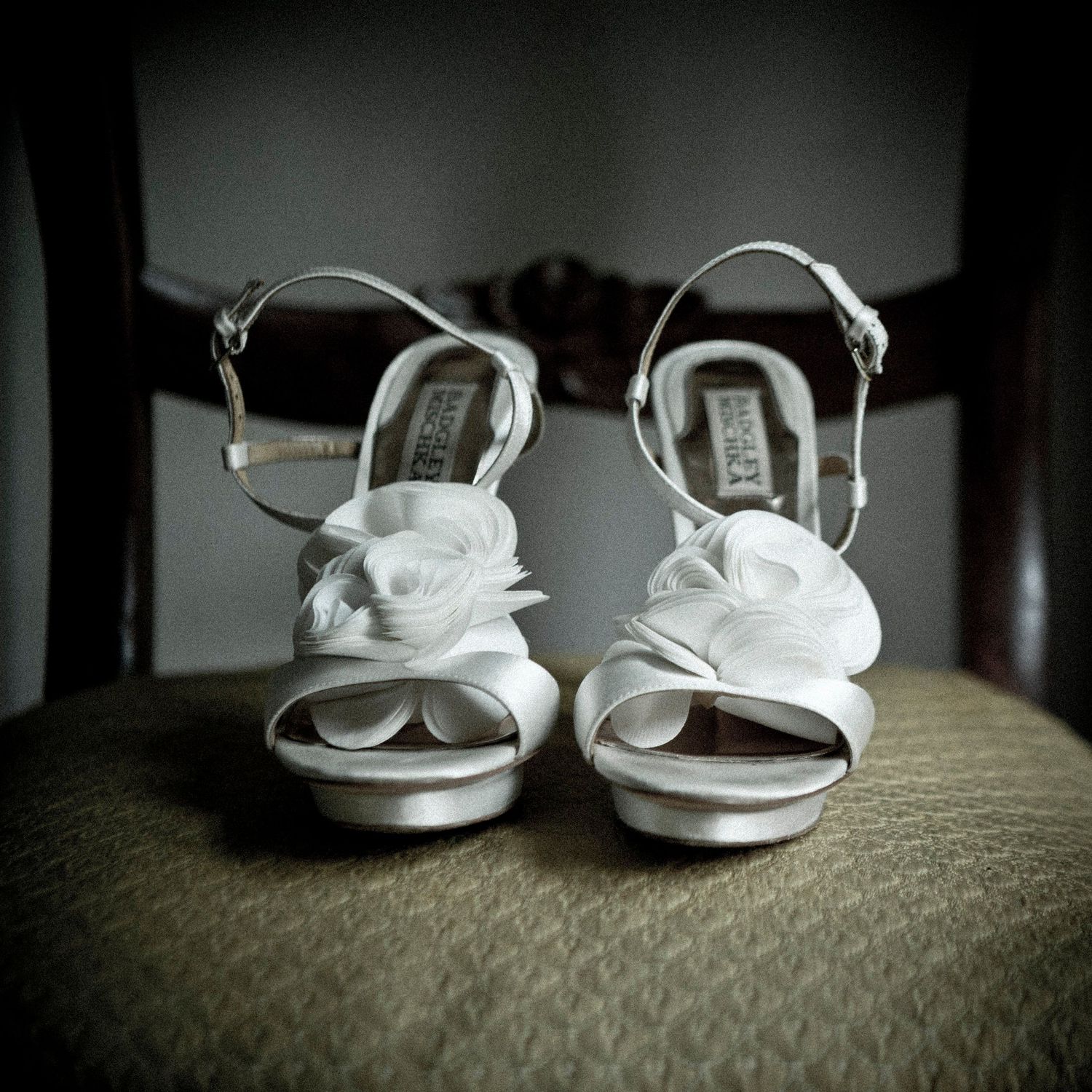 Elegant white satin bridal heels with floral rosette detail displayed on a vintage chair.
