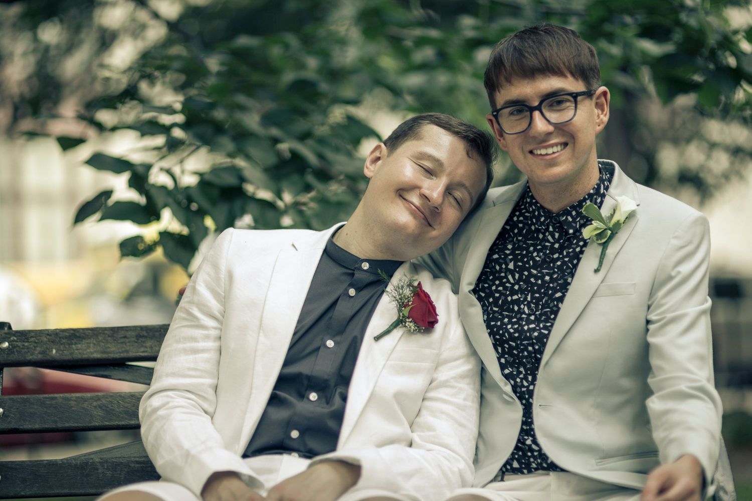 Two men in suits with boutonnieres sitting on a bench, smiling and leaning on each other outdoors.