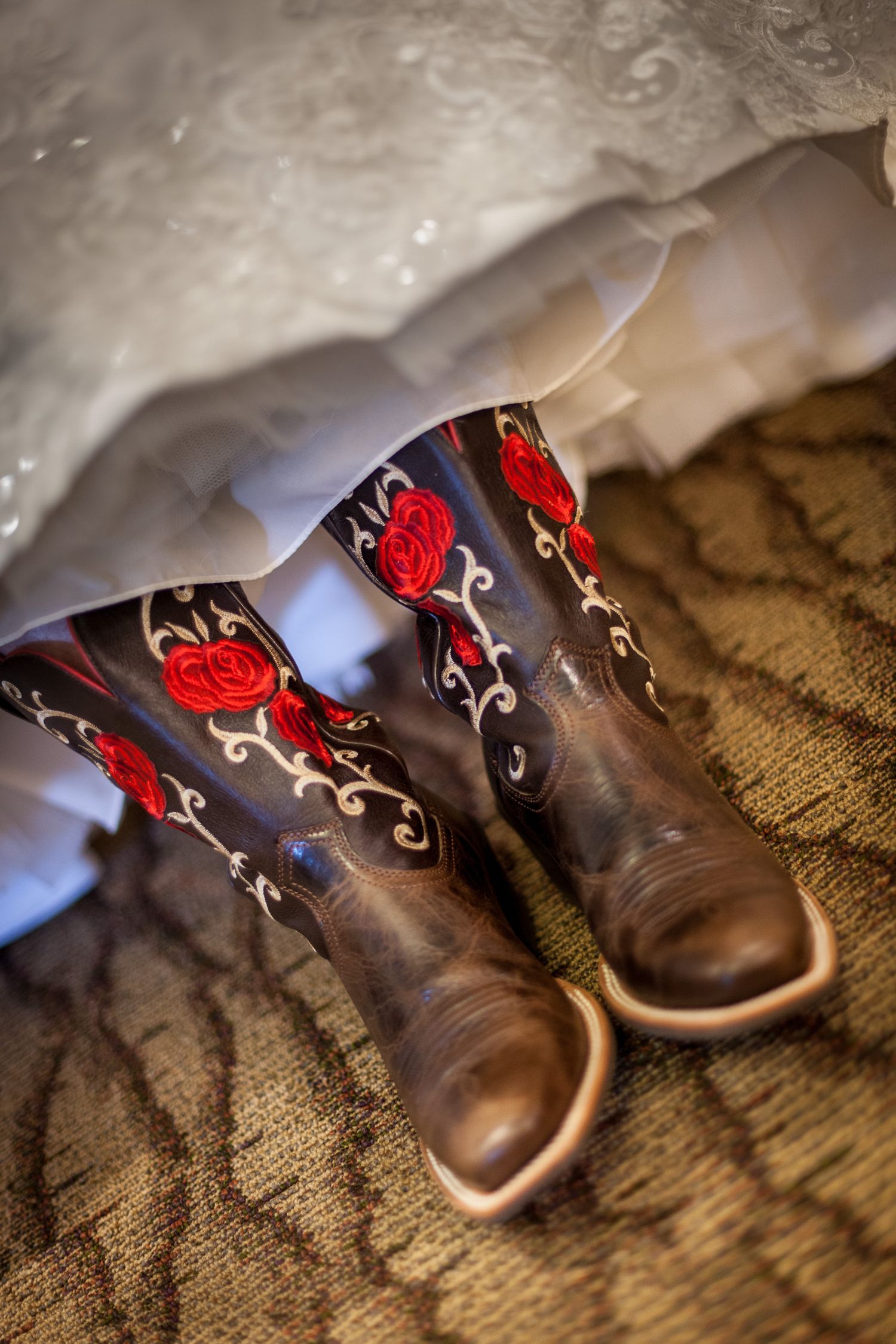 Brown cowboy boots with red rose embroidery peek beneath a white wedding dress hem on a textured carpet.