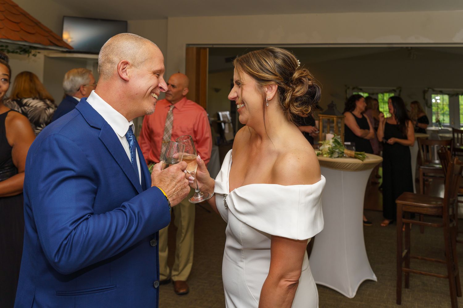 Bride and groom laughing together at wedding reception, groom in blue suit, bride in off-shoulder gown.
