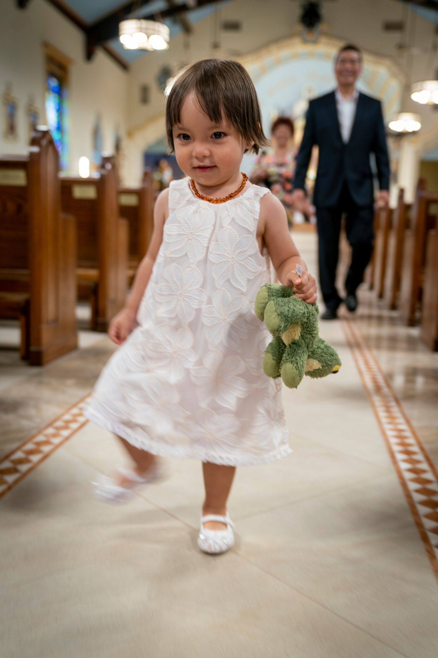 Toddler girl in white dress carrying green stuffed frog toy walking down church aisle during wedding ceremony.