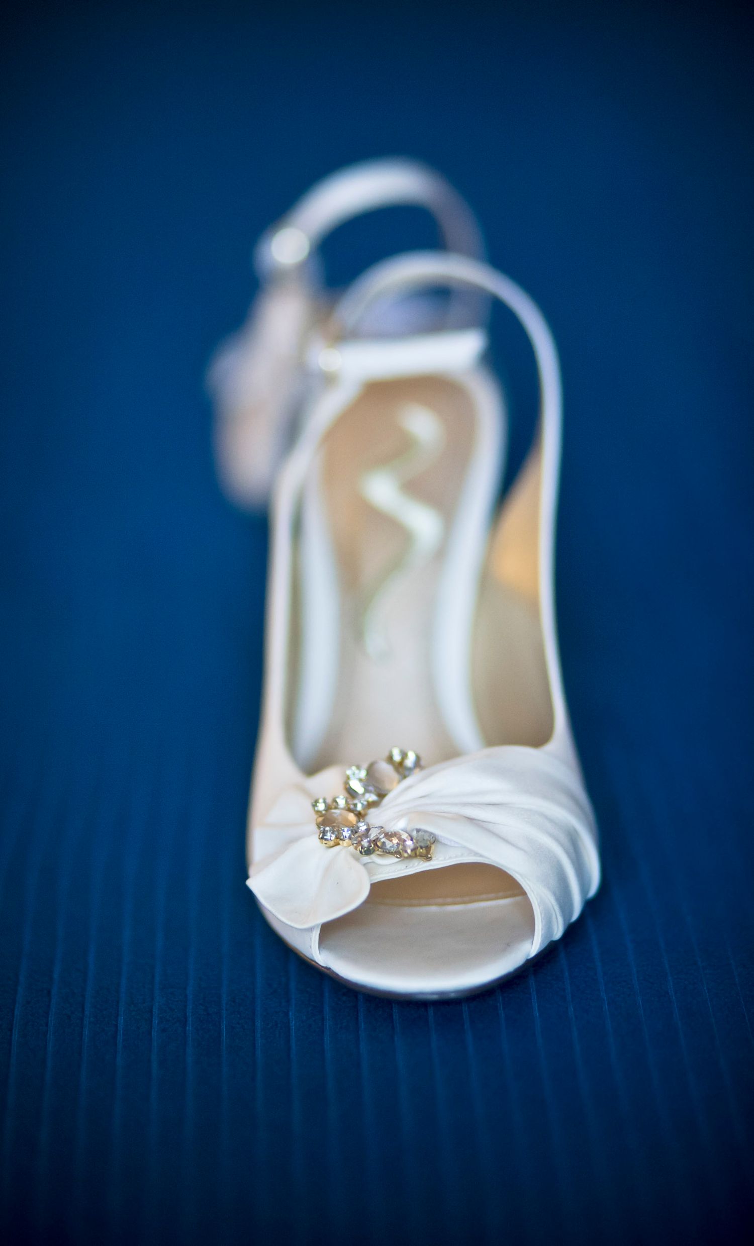 White bridal heeled sandal with pearl floral embellishment on blue textured background.