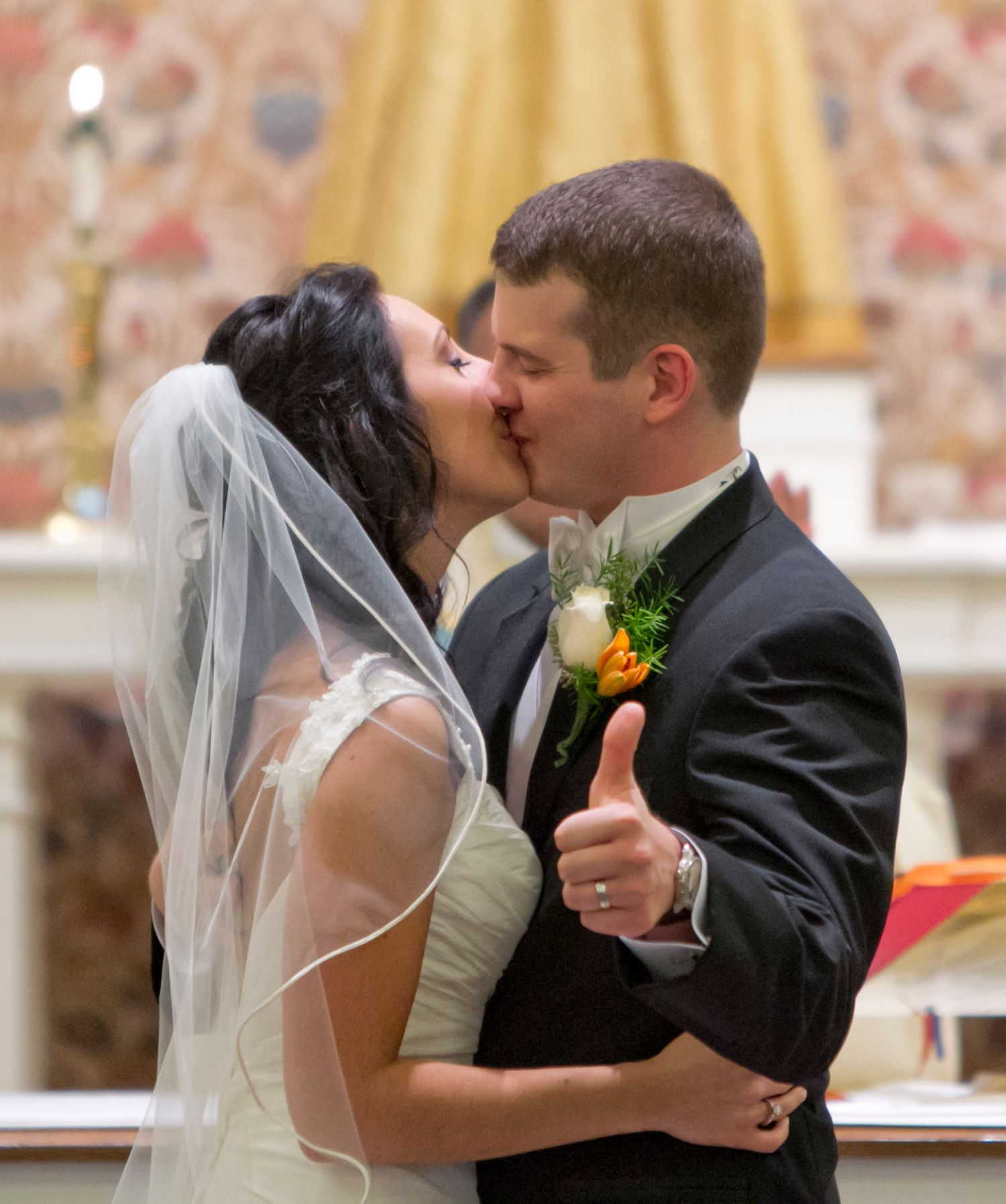 Bride and groom share first kiss at church altar, groom giving thumbs up in wedding ceremony.