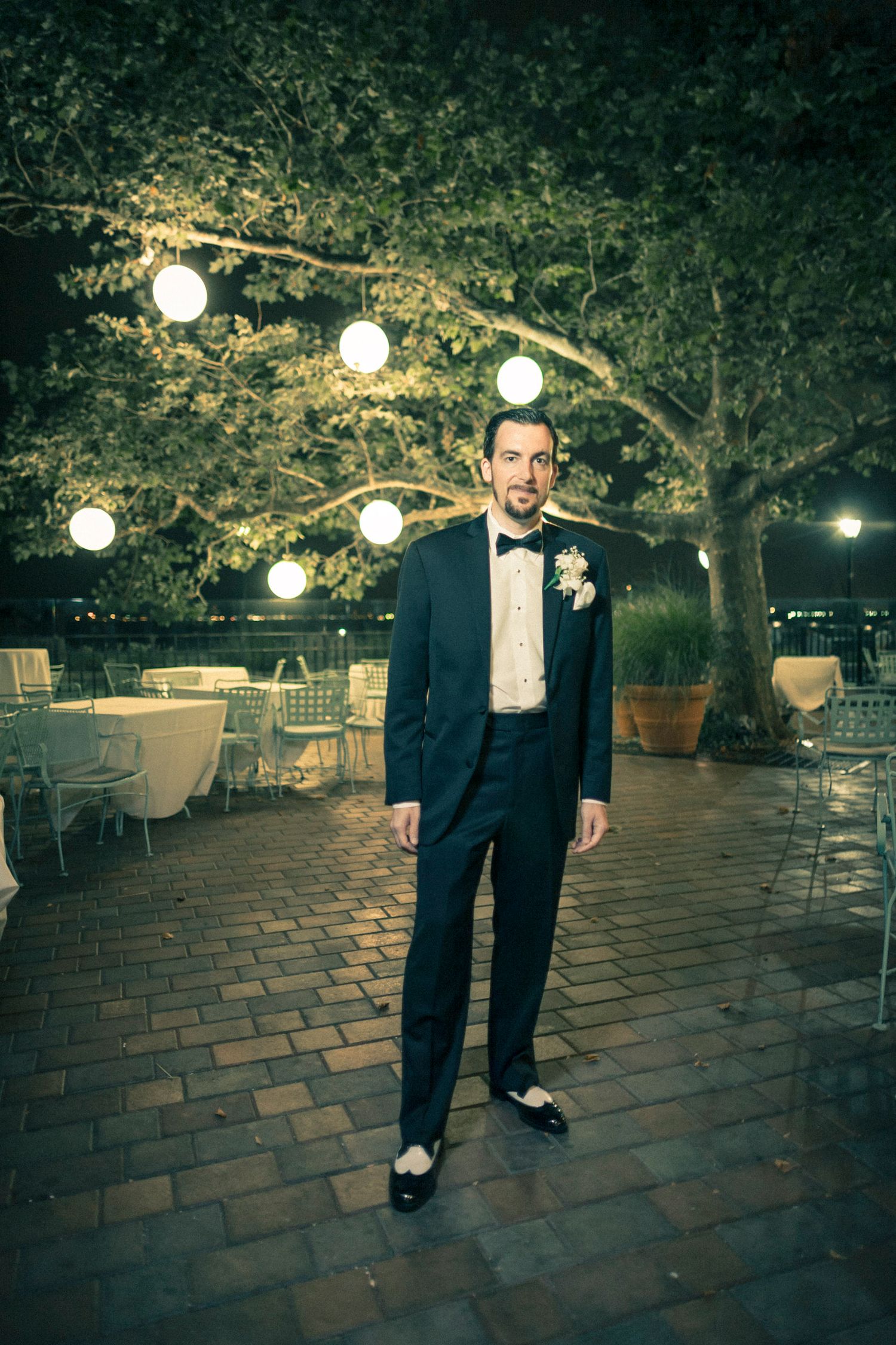 Groom in black tuxedo stands under globe-lit trees on a brick patio at night.