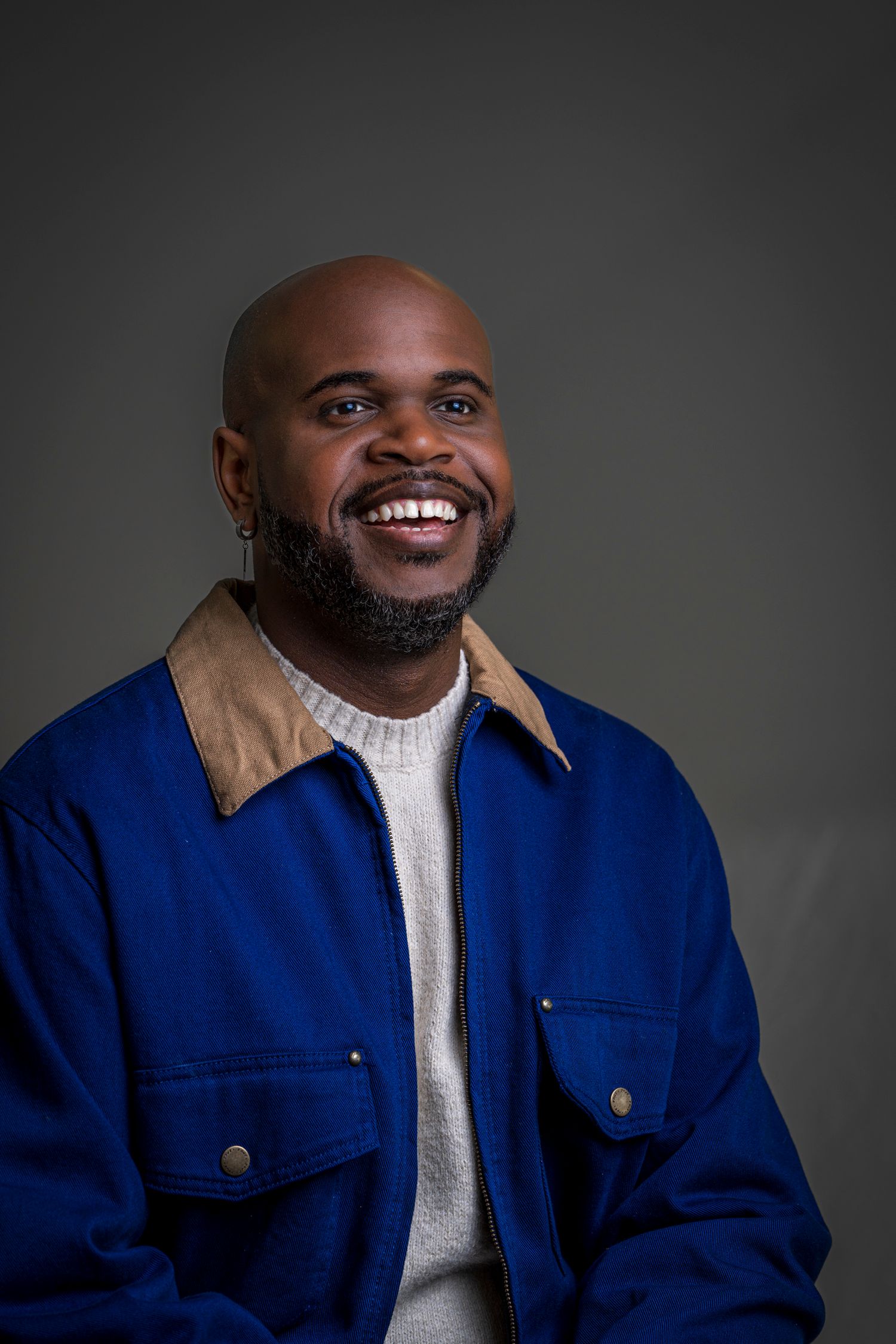 Smiling bald Black man with beard wearing a blue jacket over a gray sweater, photographed against a dark gray background.