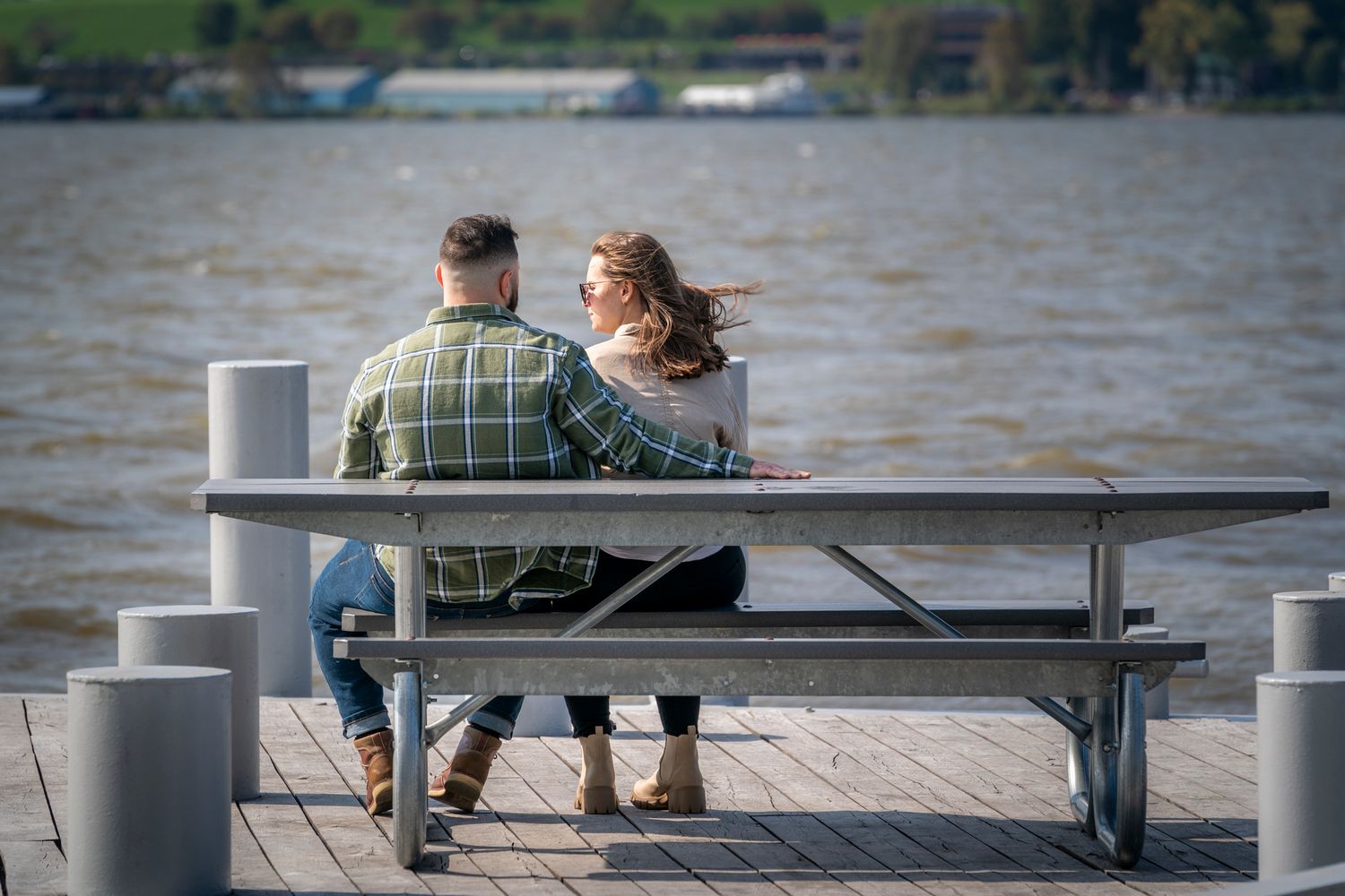 Couple sitting together at a waterfront picnic table on a wooden dock, enjoying a scenic lake view on a breezy day.