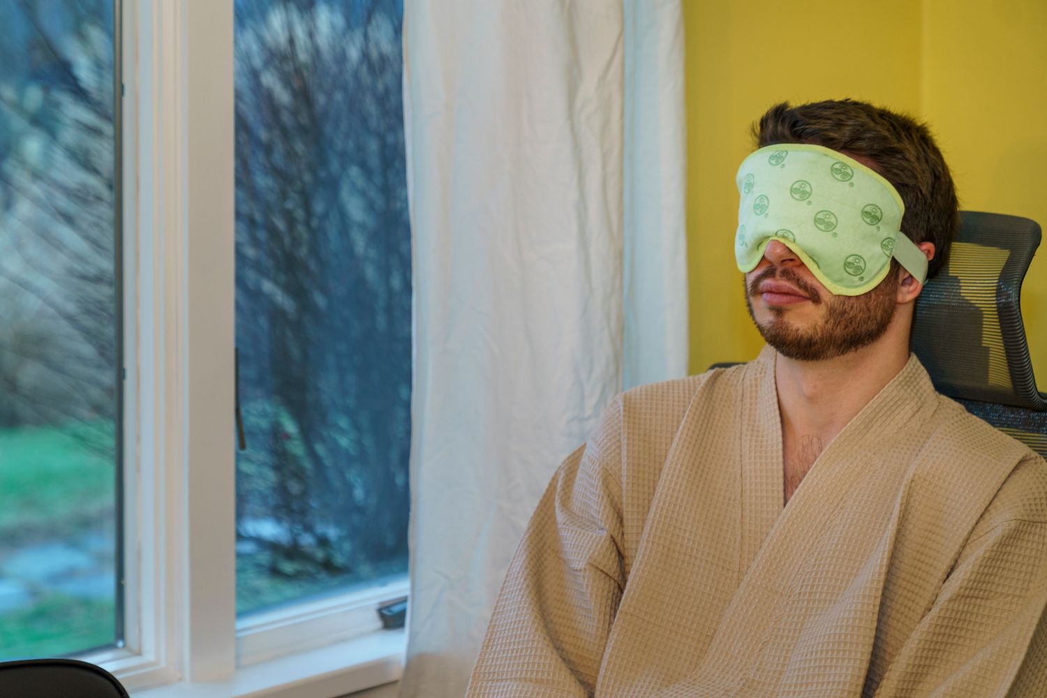 Man wearing a green sleep eye mask and beige robe relaxing in a chair near a window.