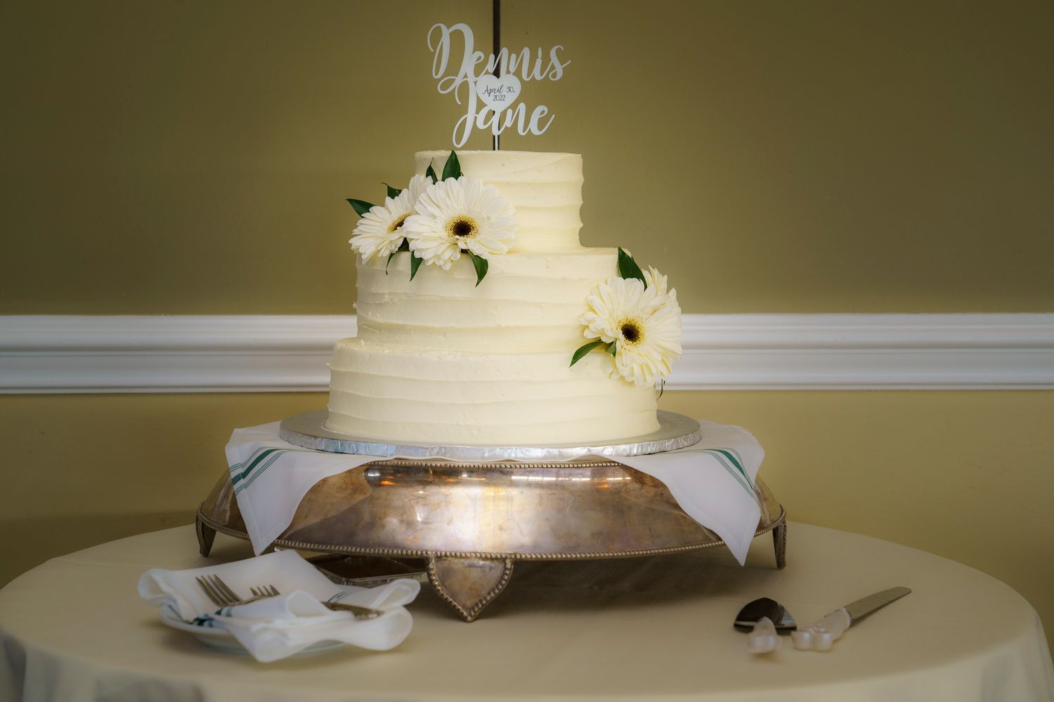 Three-tier white wedding cake with gardenia flowers and custom name topper on silver stand.