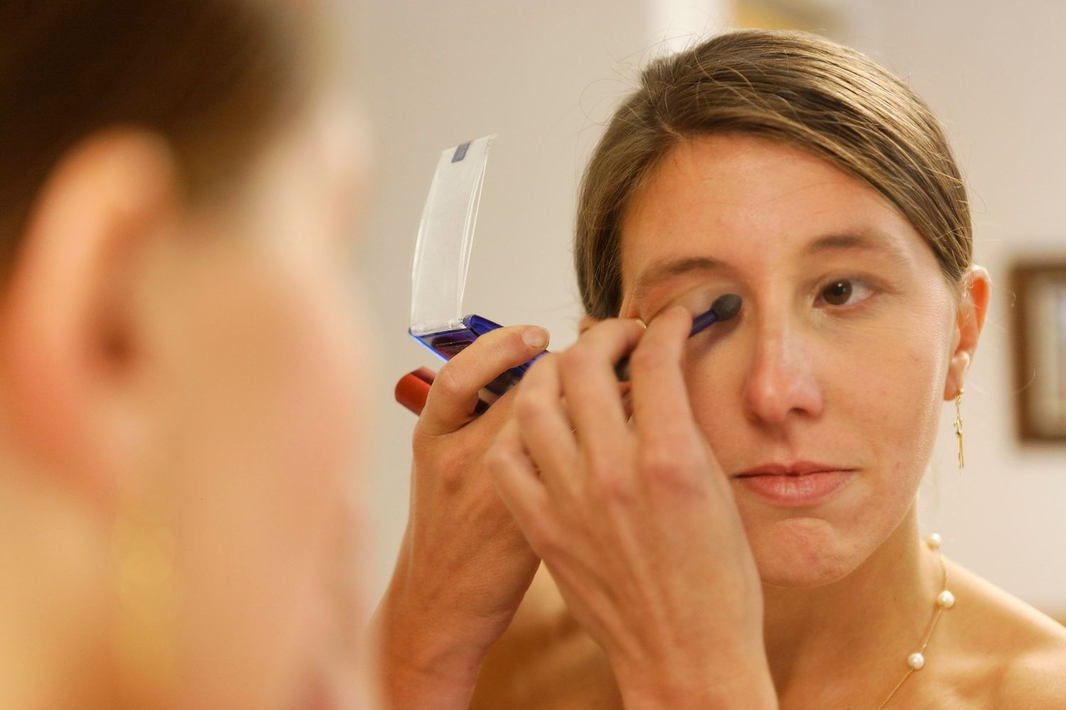 Bride applying eye makeup while looking into small mirror during wedding preparations.