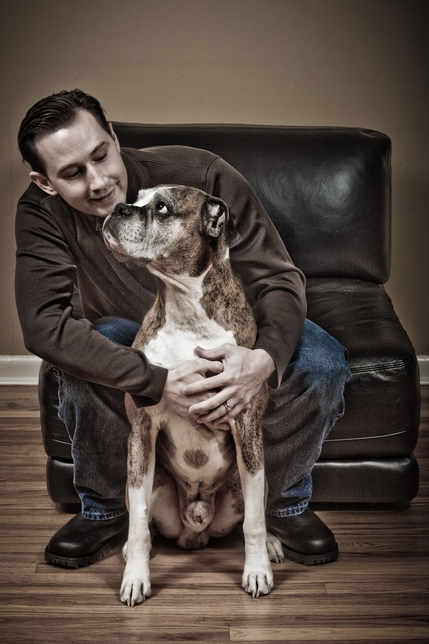 Man sitting on leather chair hugging a brindle boxer dog on hardwood floor.
