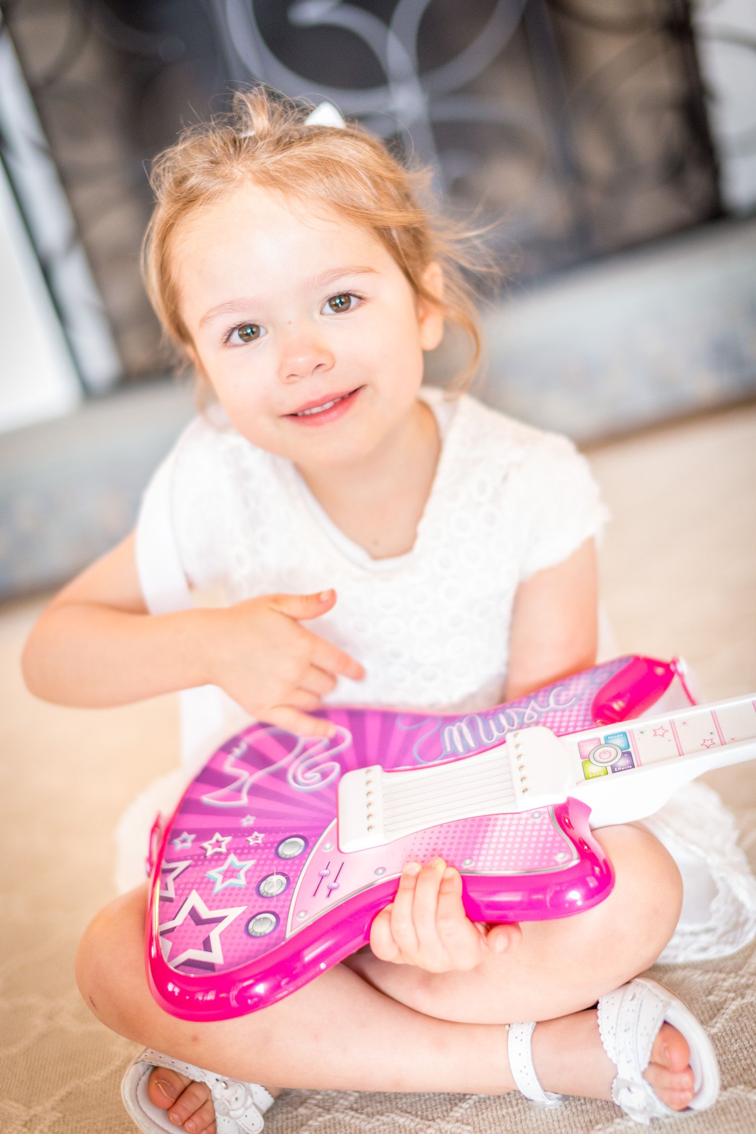 Cute toddler girl sitting on floor smiling while holding a pink toy guitar.