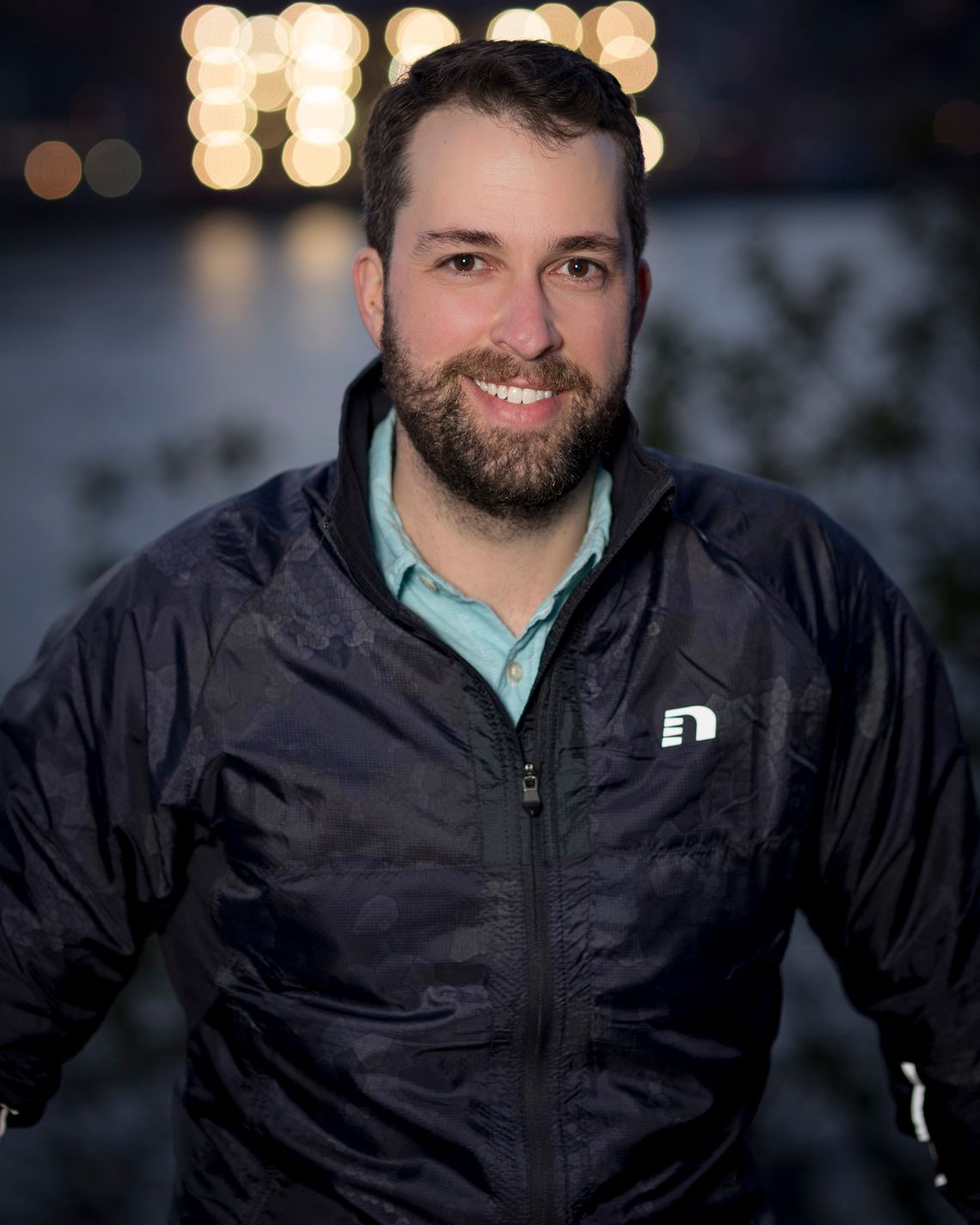 Smiling bearded man in a navy puffer jacket poses outdoors at night with bokeh city lights behind him.