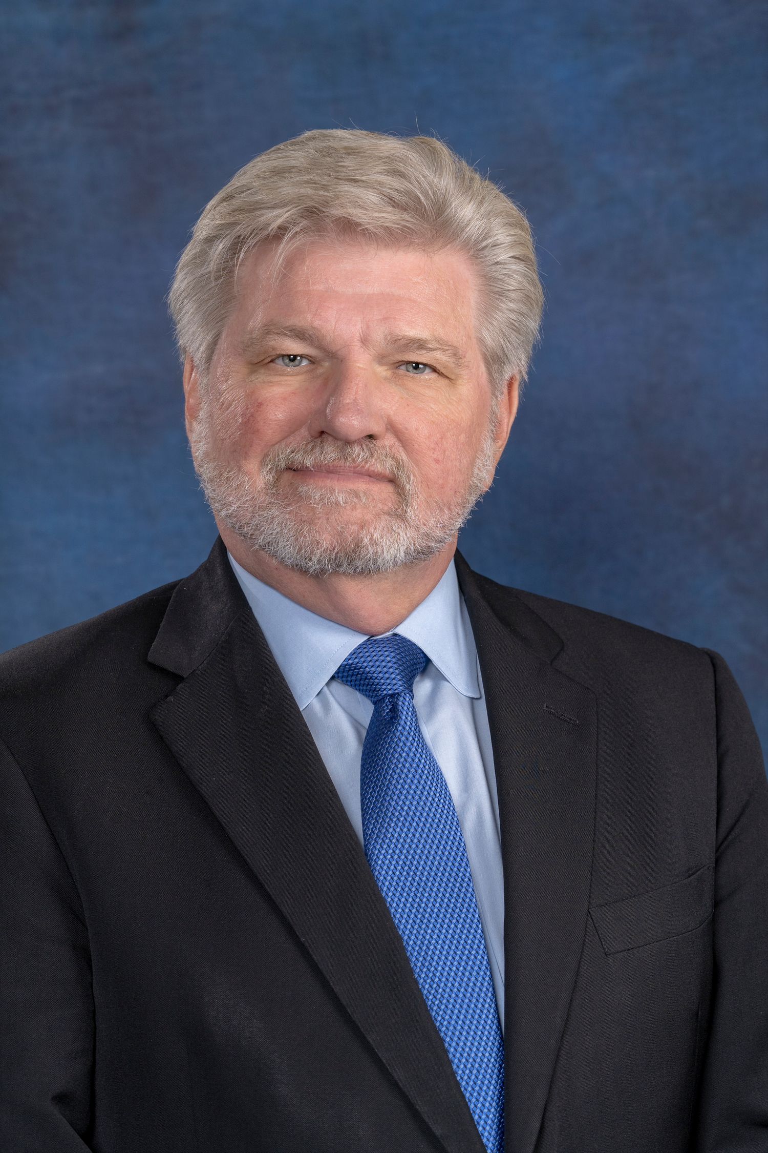 Senior businessman with gray hair and beard wearing suit and blue tie.