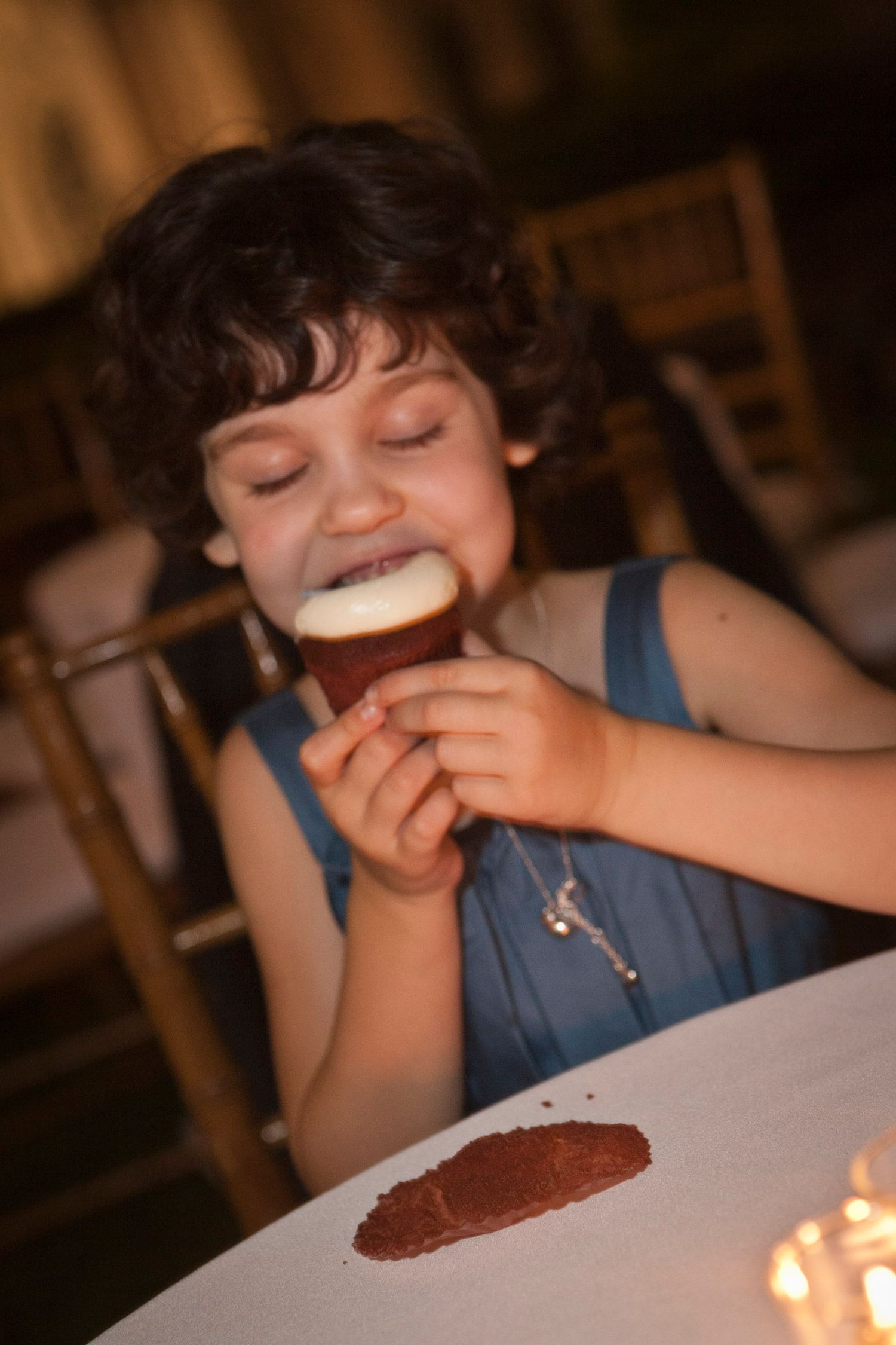 Young curly-haired girl in blue dress enjoys a frothy drink at a wedding reception table.