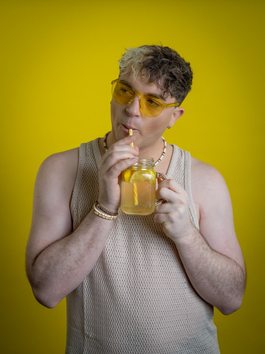 Young man in mesh tank top sipping yellow drink from mason jar, wearing yellow sunglasses against bright yellow background.