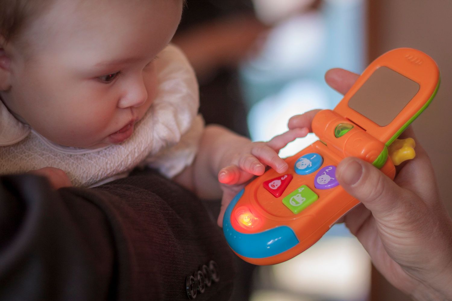 Baby reaching for a colorful orange toy flip phone held by an adult hand.