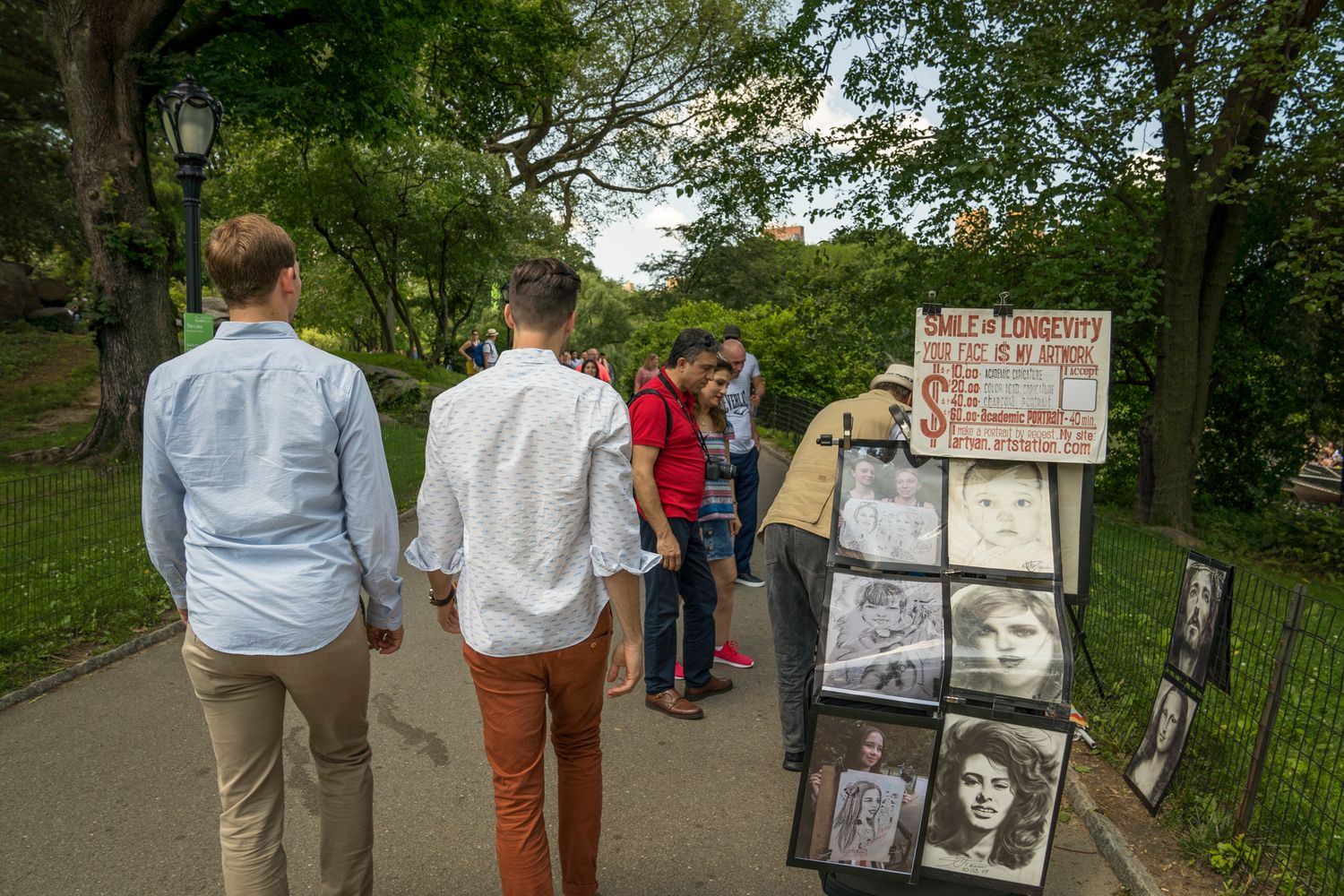 People browsing an outdoor portrait art display on a park path surrounded by lush green trees.