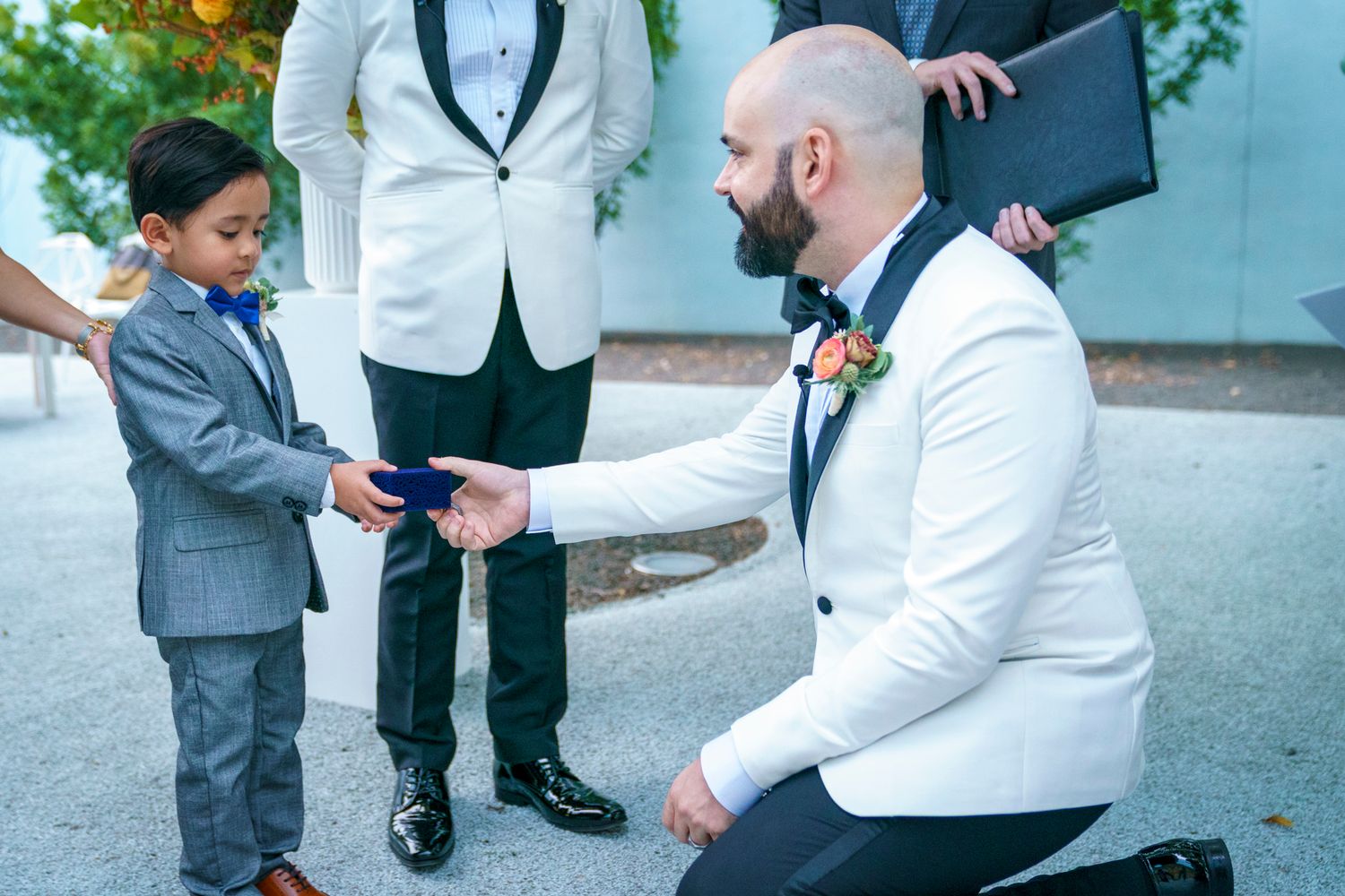 Groom kneeling to receive wedding rings from young ring bearer during outdoor ceremony.
