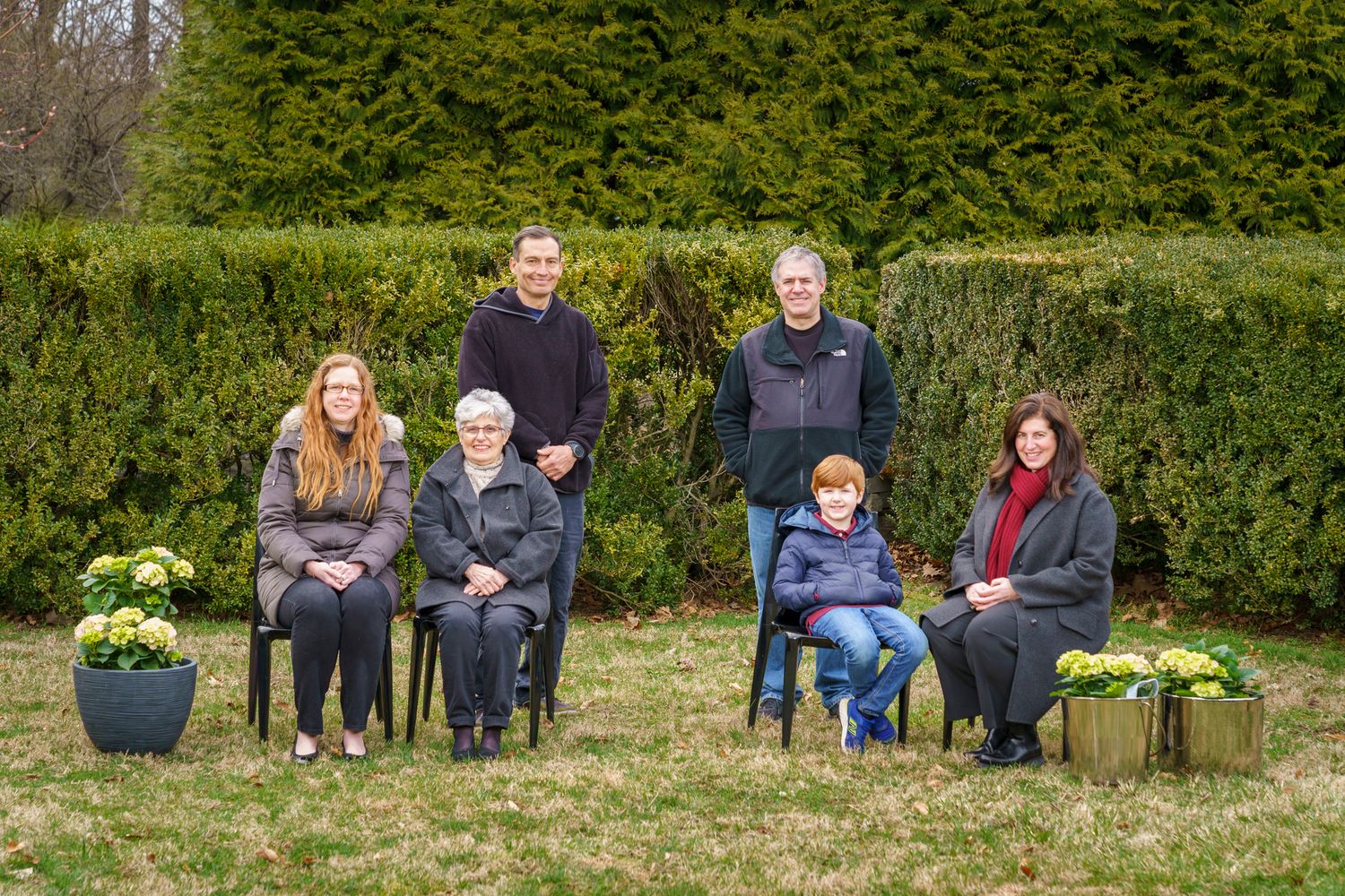 Family group portrait outdoors with hedgerow backdrop, two standing men and four seated members on chairs in garden.