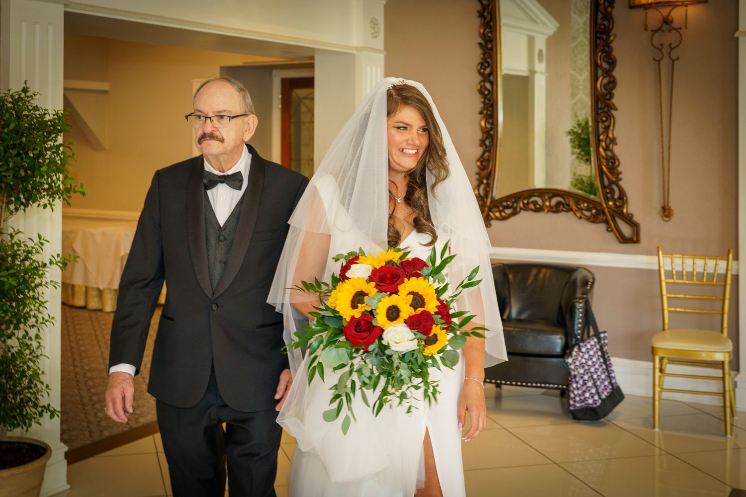 Bride in white gown and veil holding sunflower bouquet walks with suited man at indoor wedding ceremony.