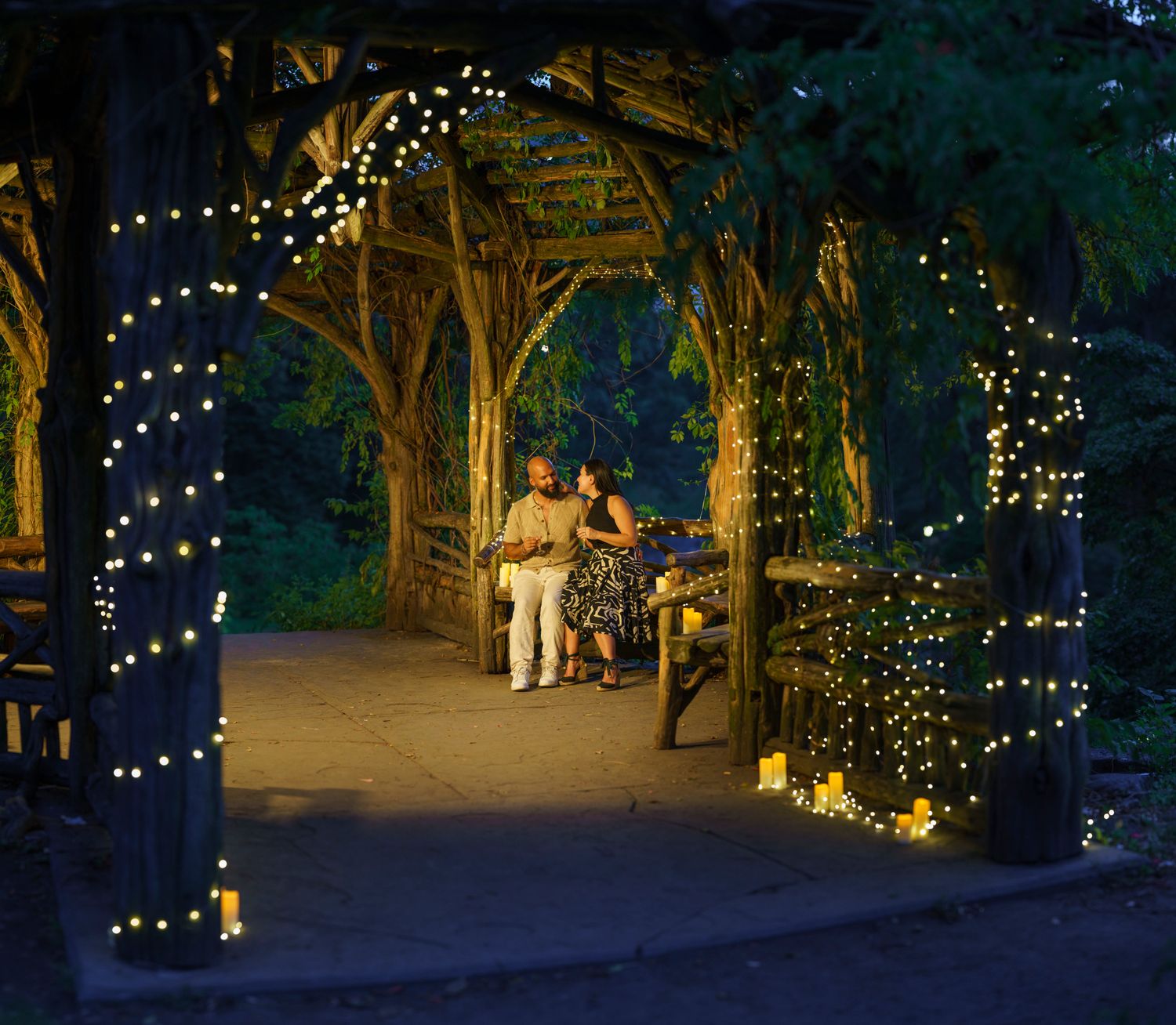 Couple sitting on a bench under a wooden pergola adorned with warm fairy lights in a romantic garden at night.