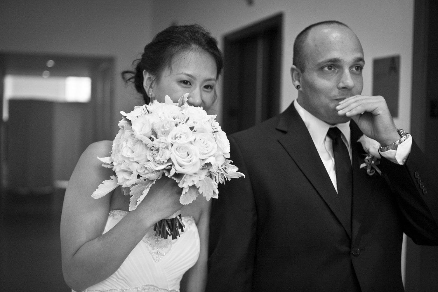 Emotional bride smelling white rose bouquet beside groom in black suit at wedding ceremony.