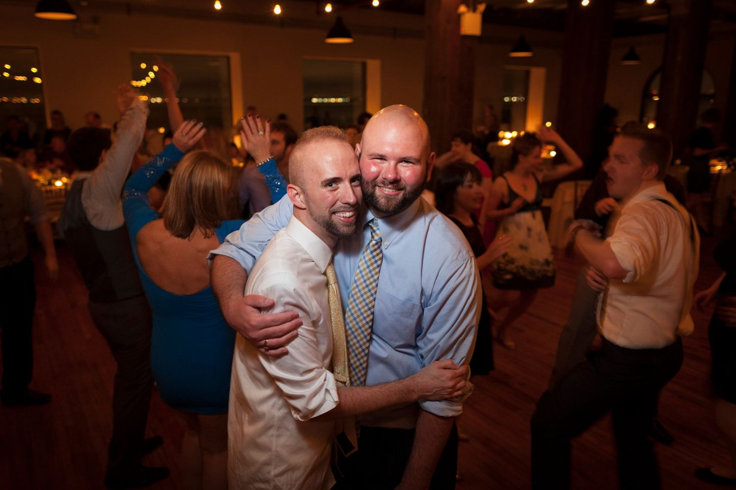 Two men share a joyful embrace on a crowded wedding reception dance floor with guests celebrating behind them.