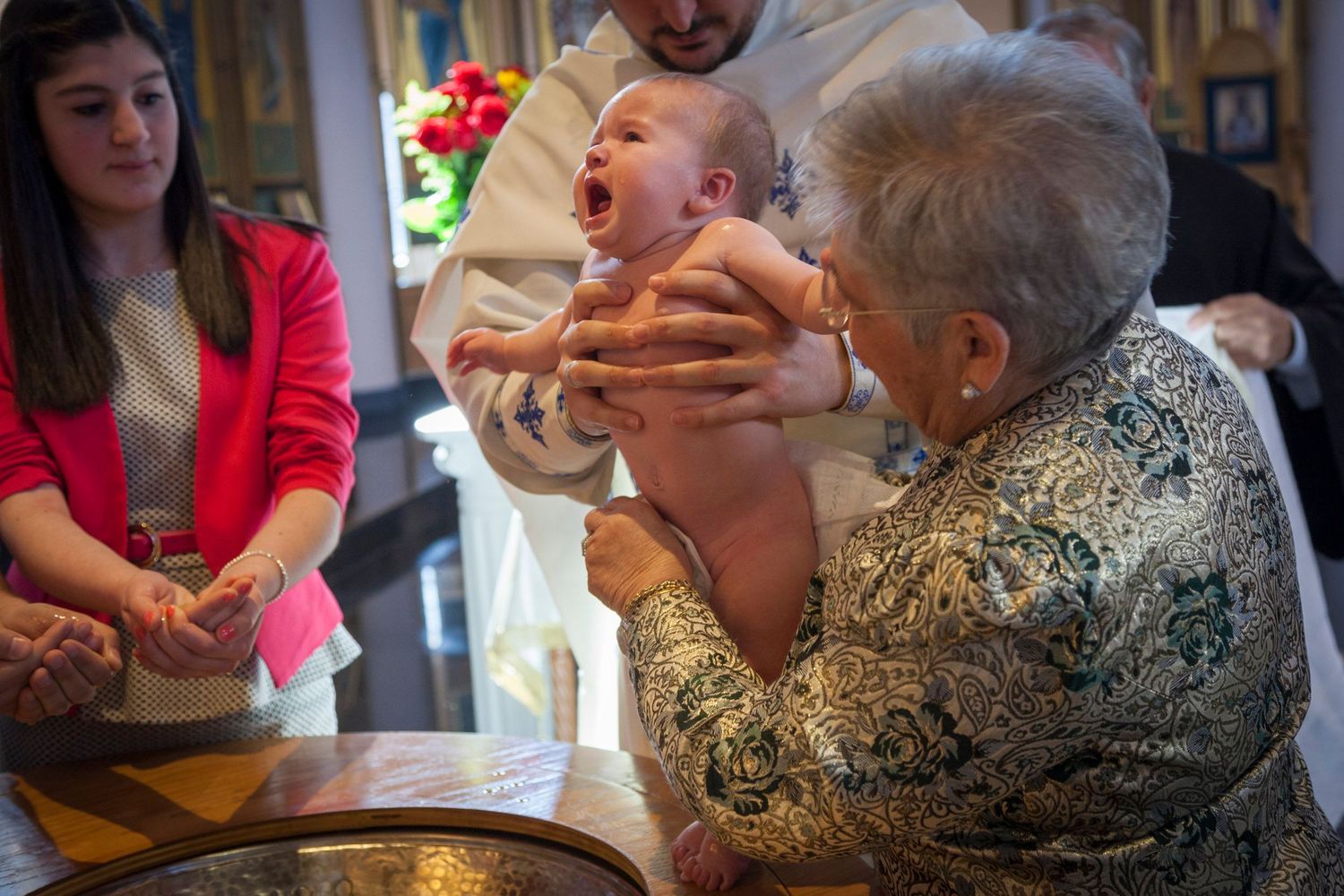 An elderly woman holds a crying baby during a Christian baptism ceremony at a church font.