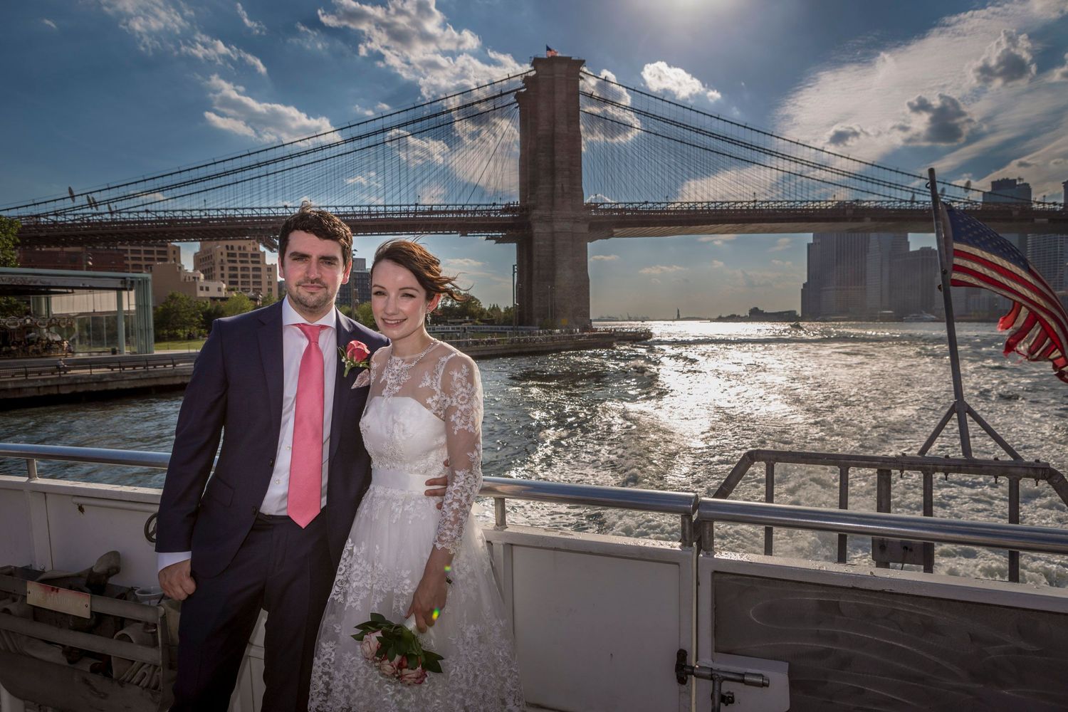 Bride and groom posing on a boat deck with the Brooklyn Bridge and East River in the background on a sunny day.