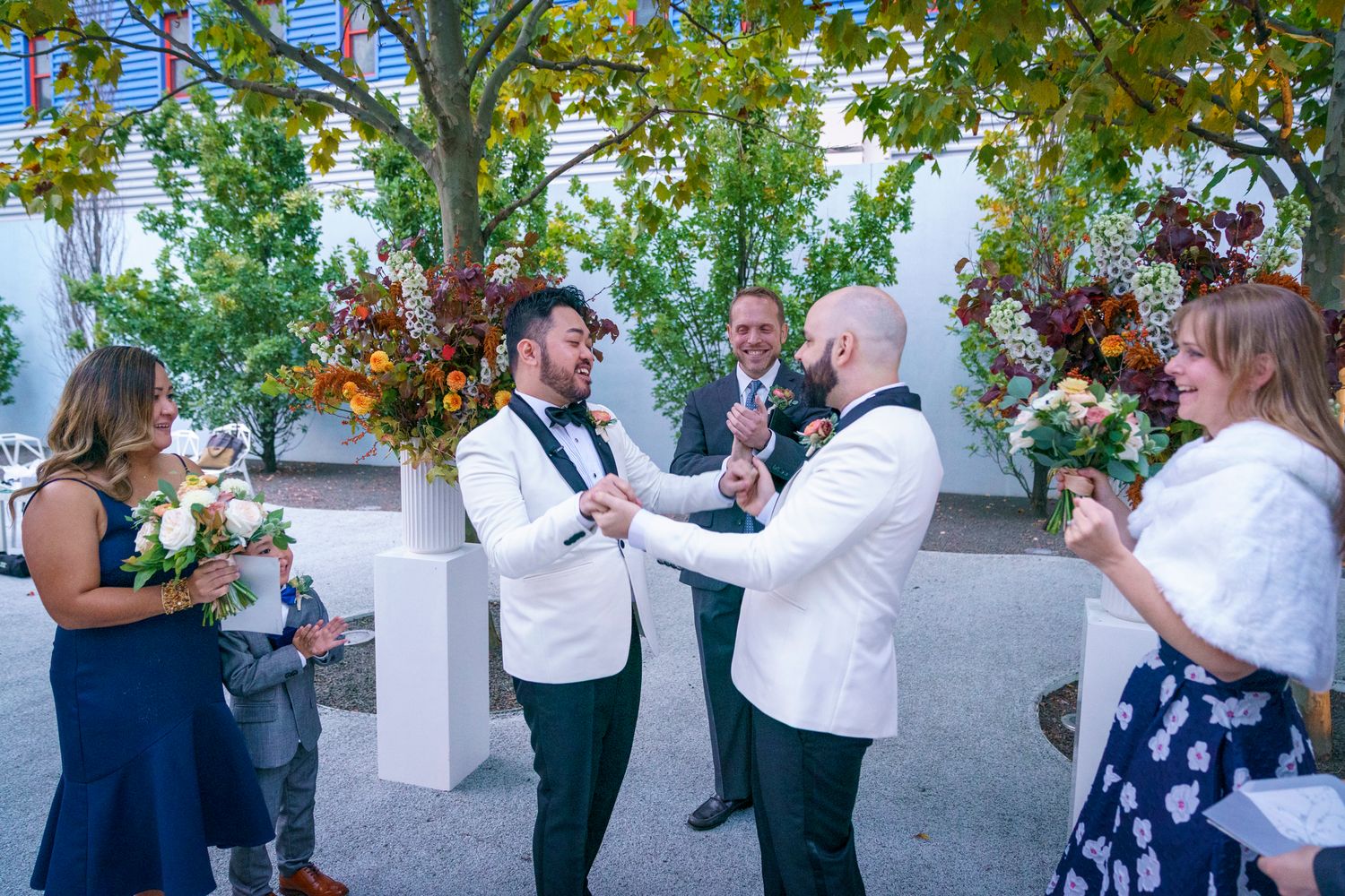 Two grooms in white tuxedos exchange vows outdoors surrounded by guests and colorful floral arrangements.