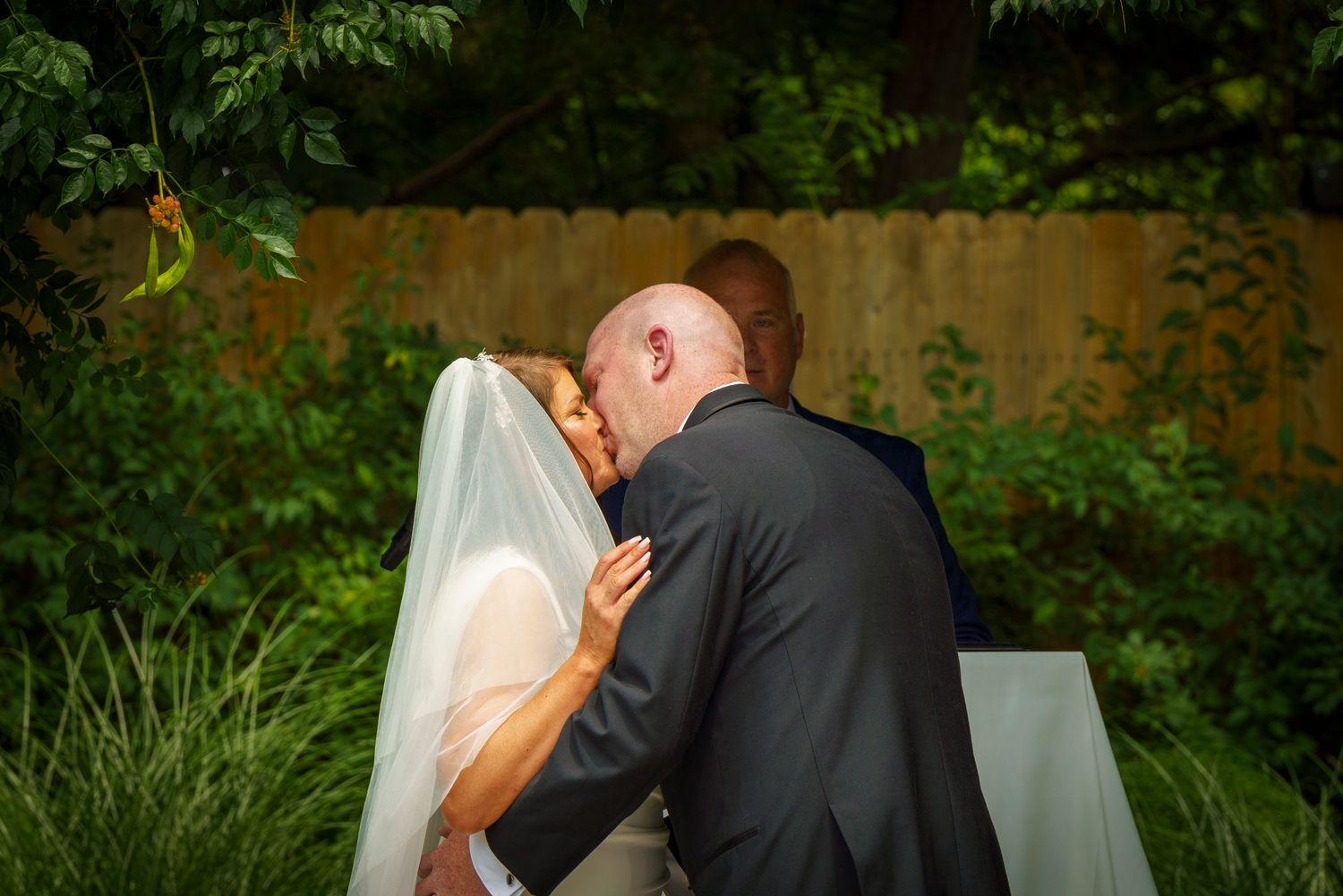 Bride and groom sharing their first kiss at an outdoor wedding ceremony surrounded by lush green foliage.