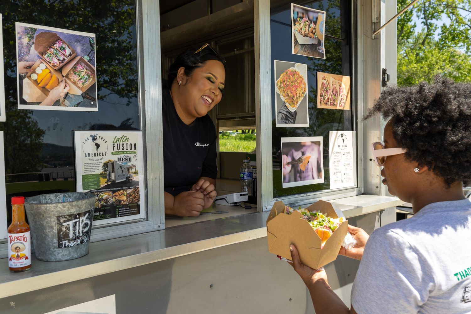 A smiling food truck vendor hands a colorful dish to a happy customer through the service window on a sunny day.