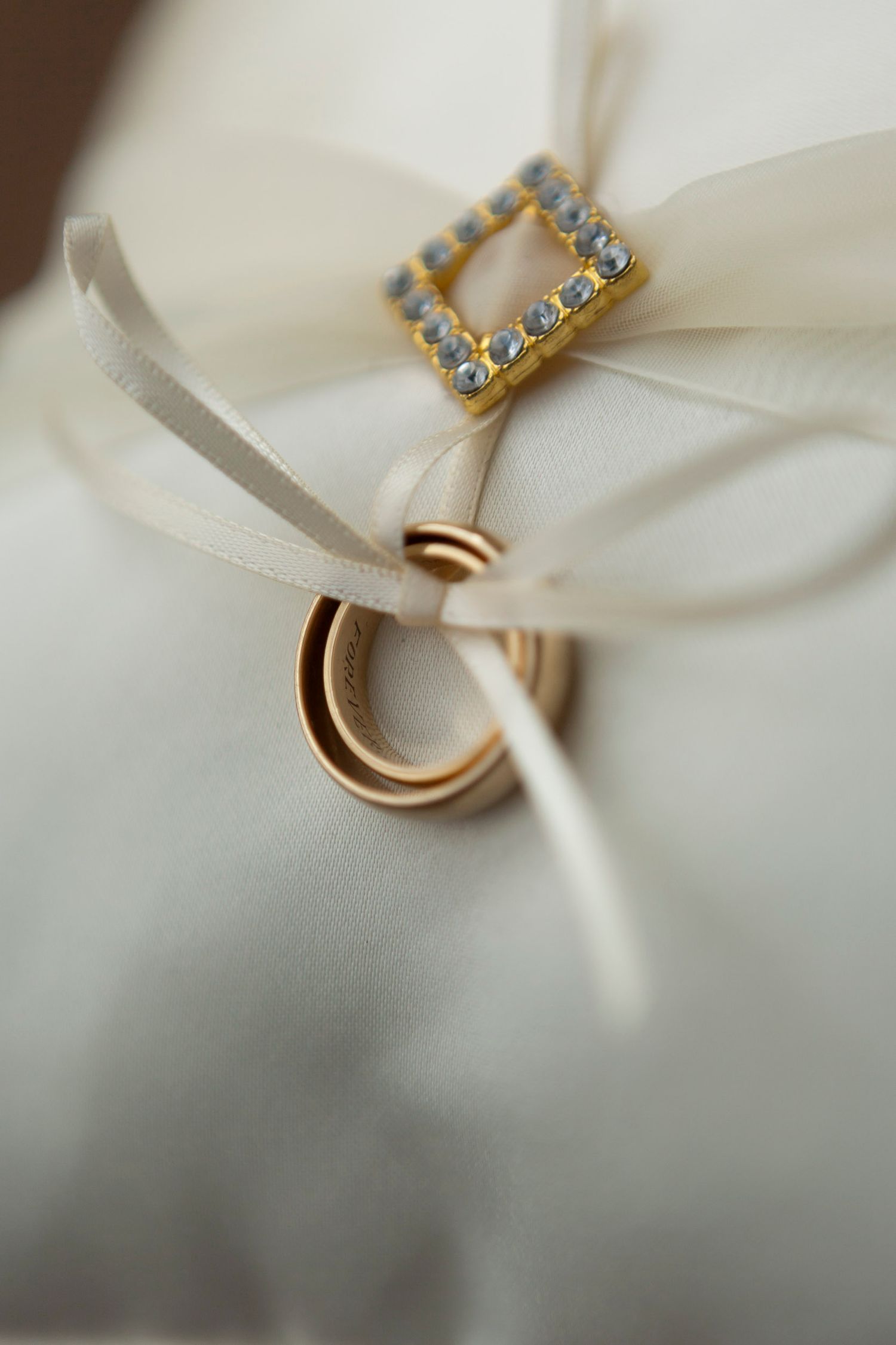 Two gold wedding bands tied with ivory ribbon on a white ring bearer pillow with diamond buckle accent.
