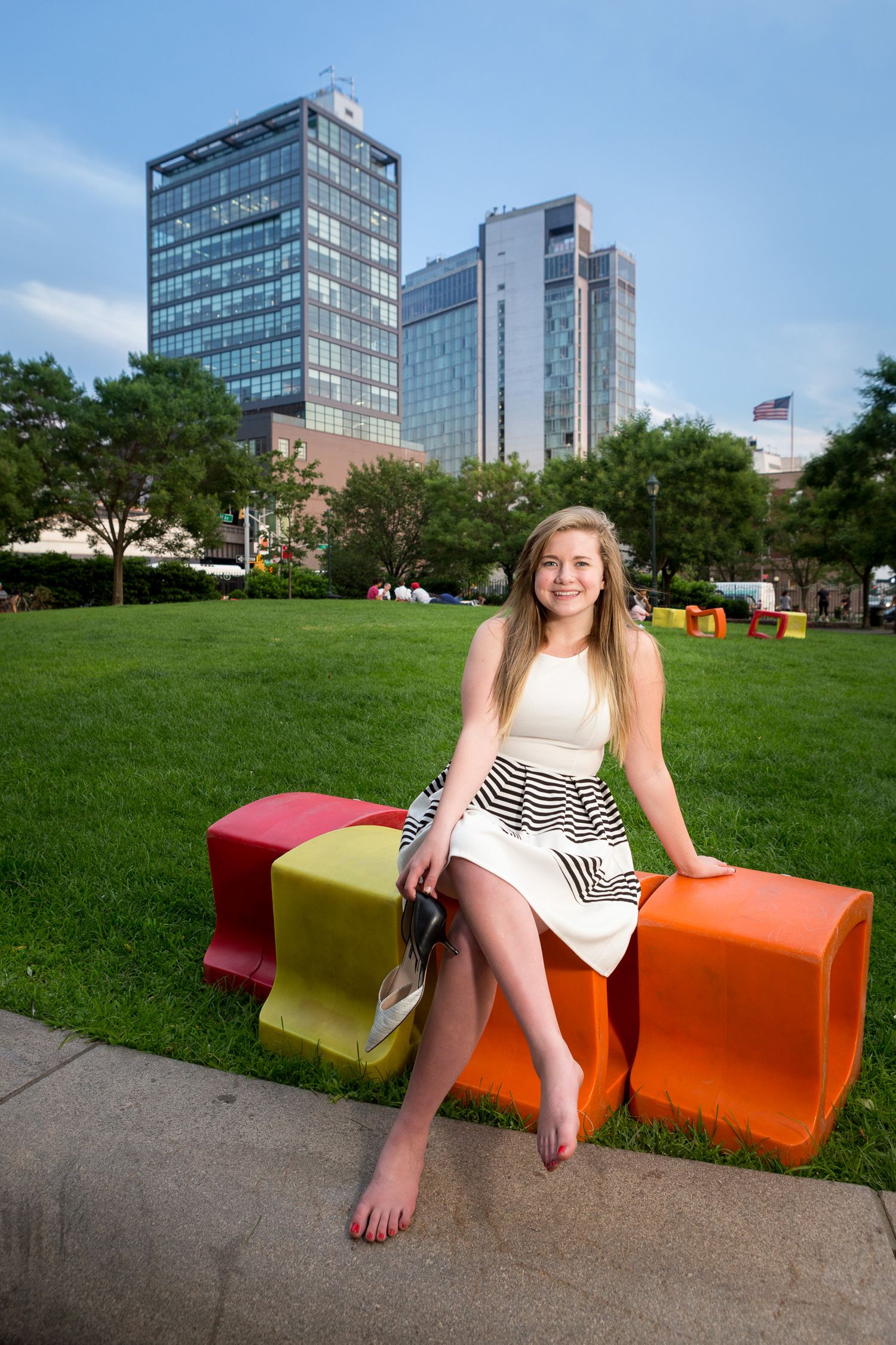 Young blonde woman in white dress sitting on colorful benches in urban park.