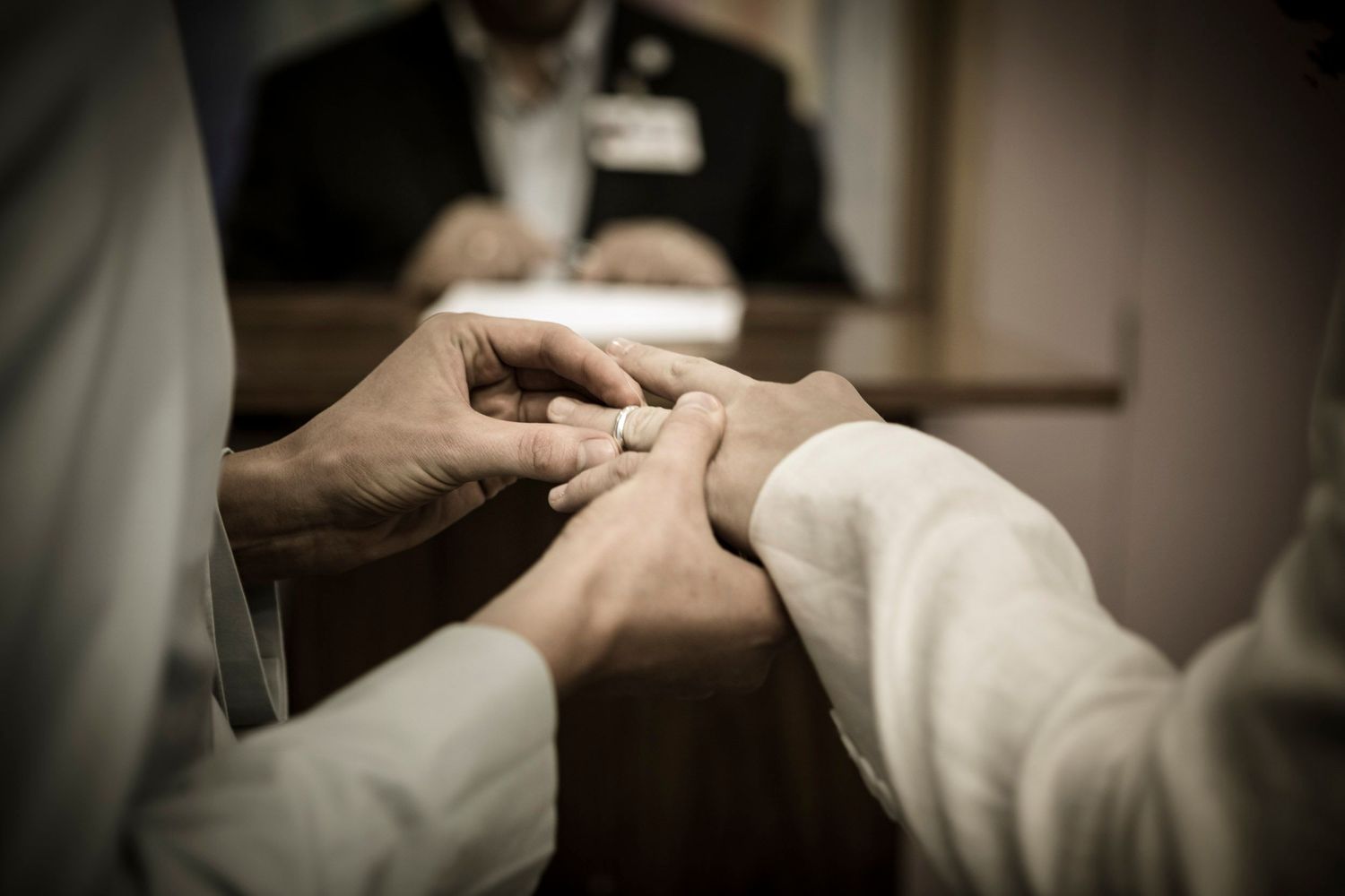 Groom placing a wedding ring on bride's finger during an intimate ceremony, with officiant visible in background.