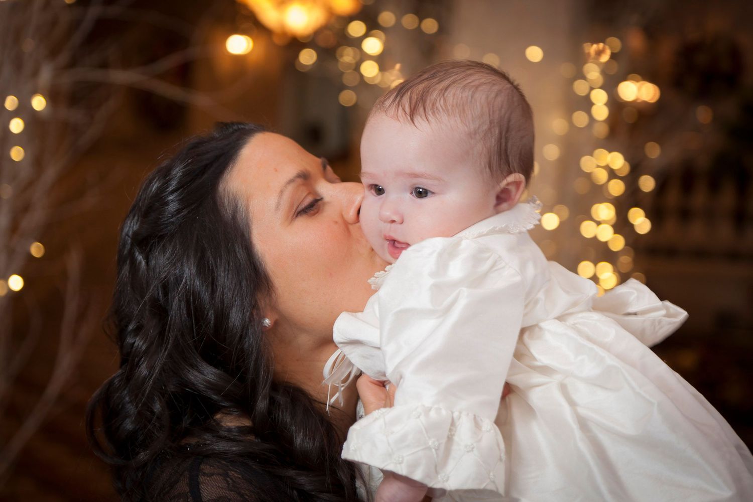 Mother tenderly kissing her baby dressed in white christening gown with bokeh lights background.