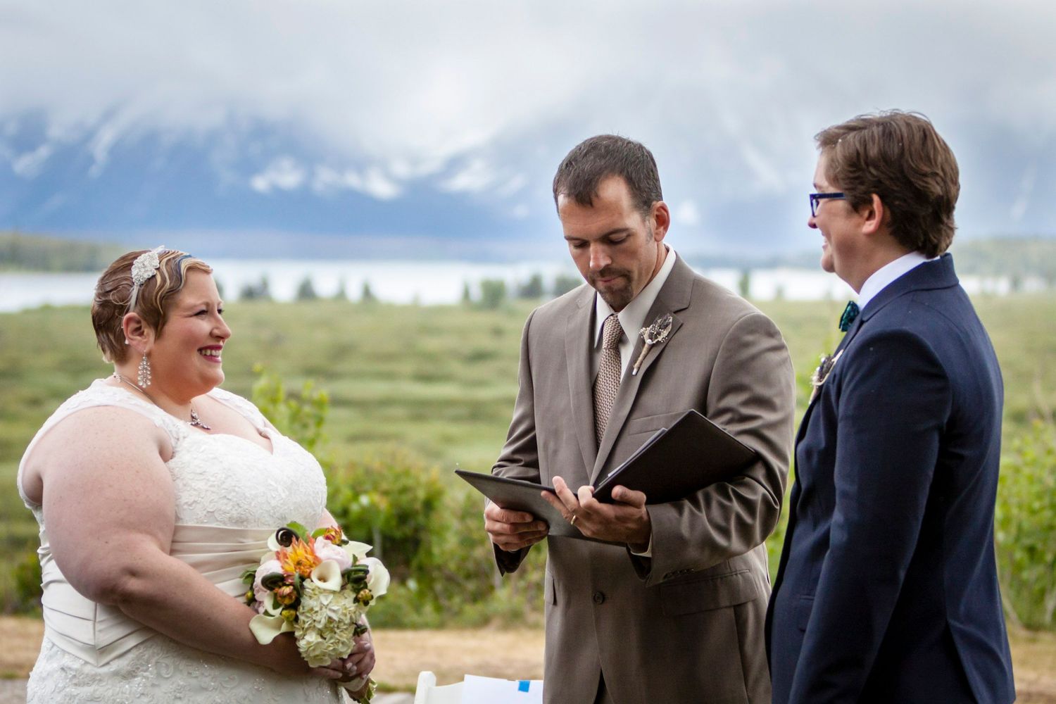 Outdoor wedding ceremony with bride, groom, and officiant against a stunning mountain lake backdrop.
