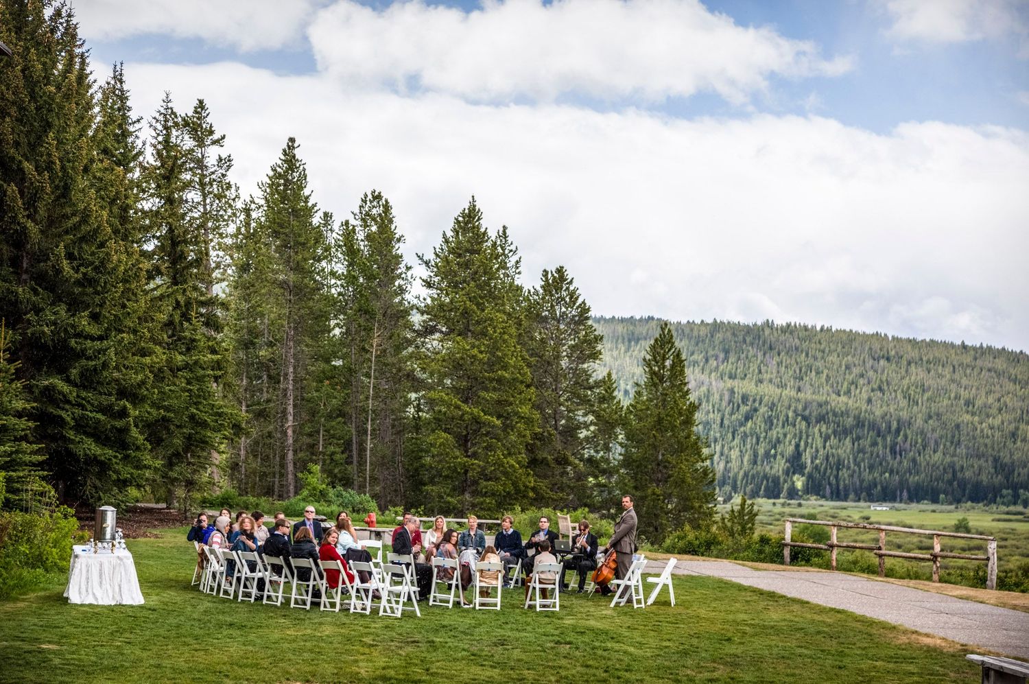Outdoor wedding ceremony with white chairs on green lawn surrounded by tall pine trees and mountain forest backdrop.