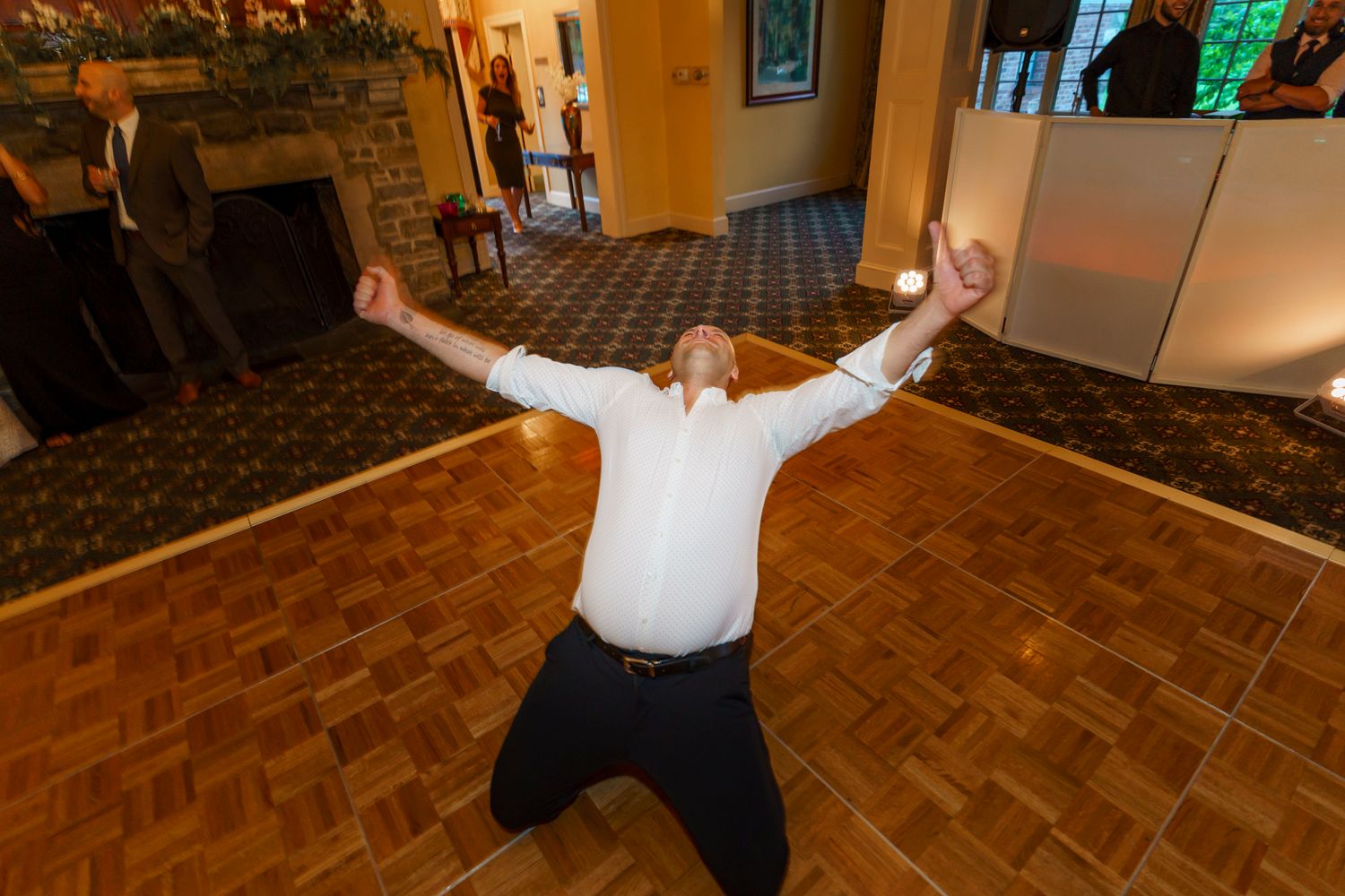 Man kneeling on dance floor with arms raised at elegant indoor wedding reception near fireplace.