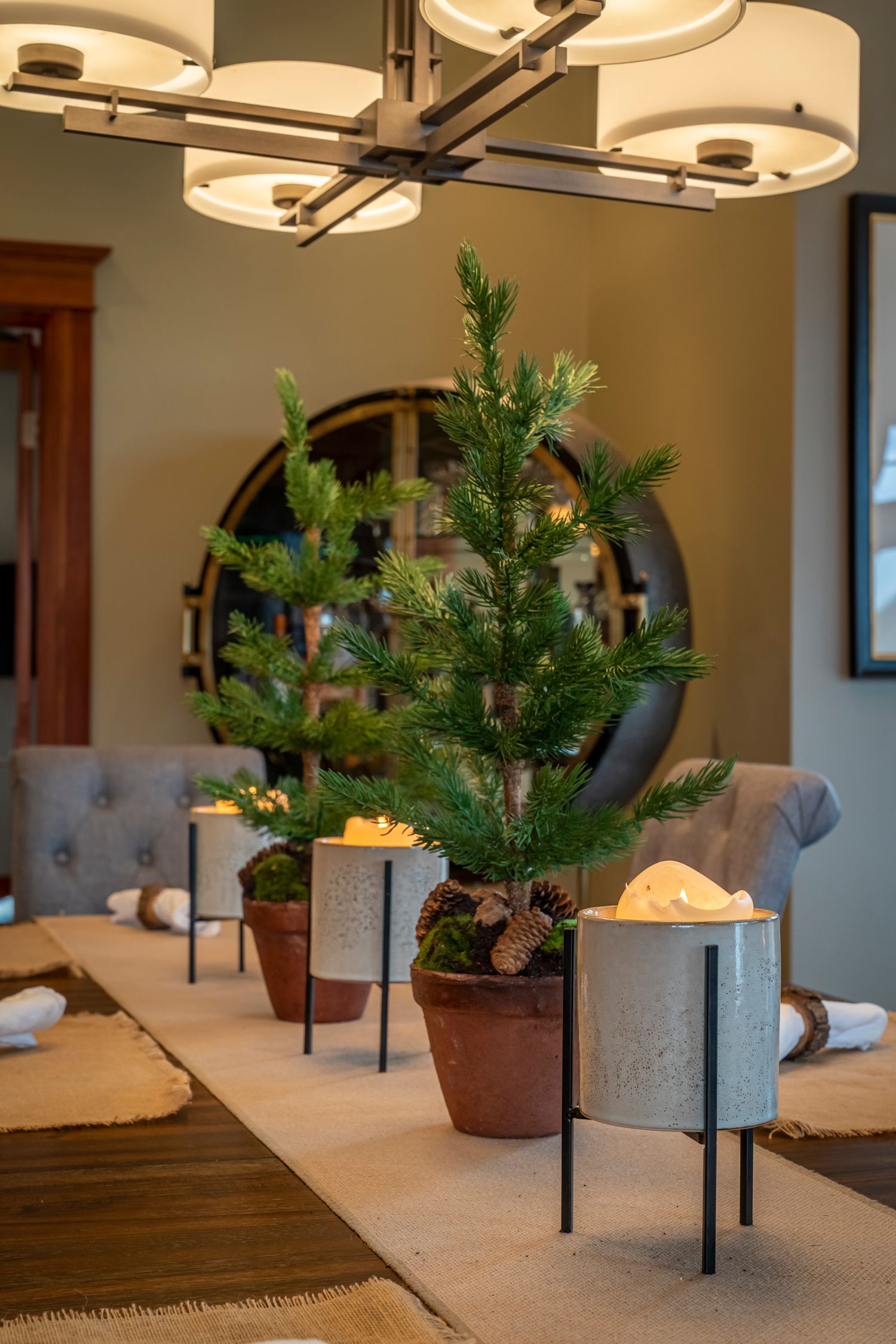 Elegant dining table centerpiece with small pine trees in metal stands, pinecones, and candles under a modern chandelier.