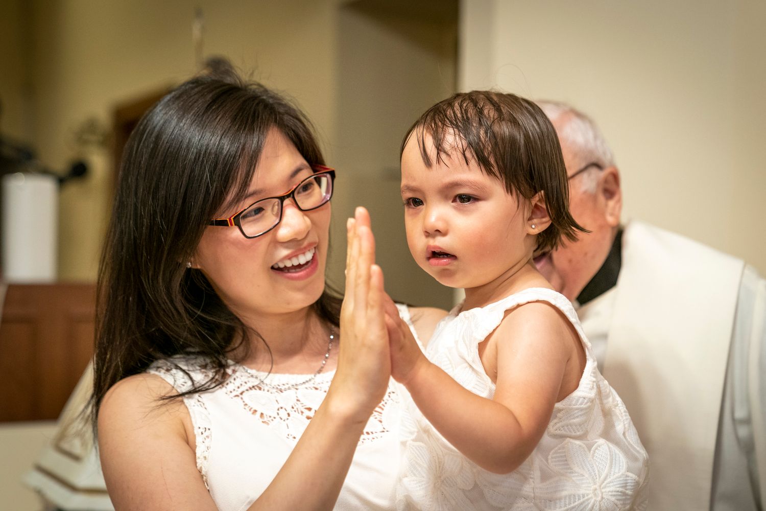 Woman with glasses playing high five with a toddler girl in white dress at a baptism ceremony.