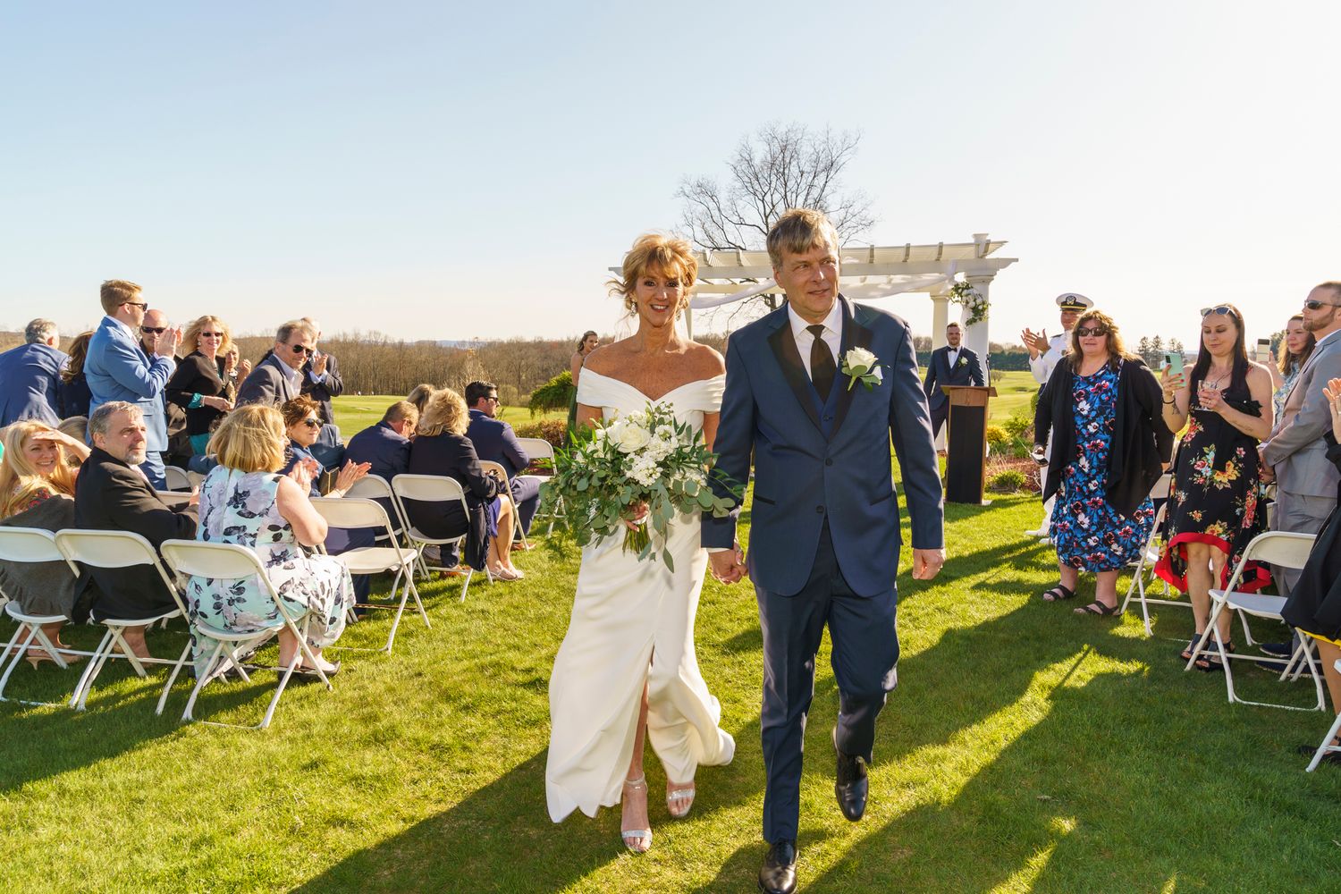 Bride and groom walk down outdoor aisle after wedding ceremony on sunny day with guests seated on white chairs.