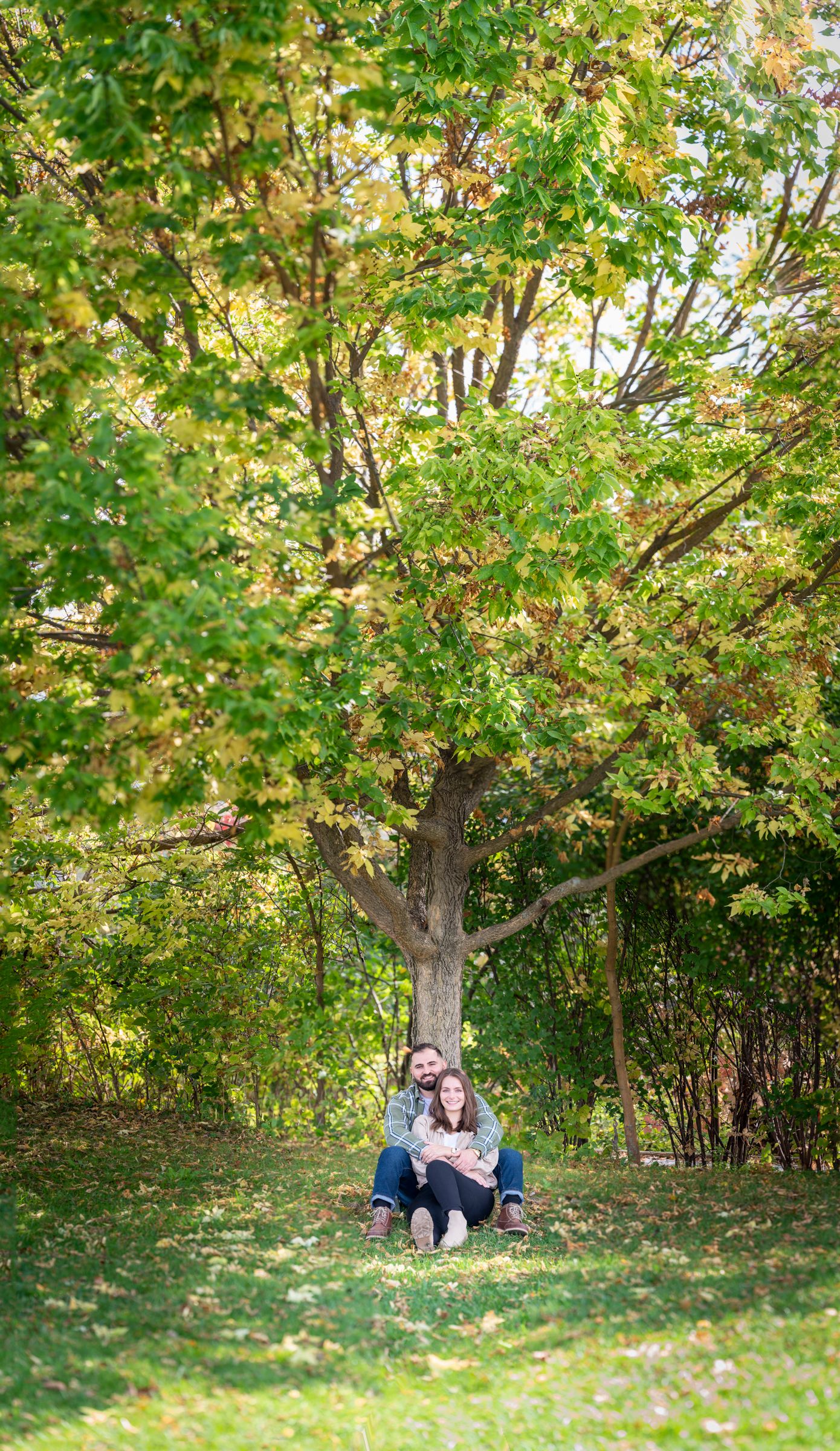 Couple sitting together under a large tree on green grass surrounded by lush foliage during autumn season.