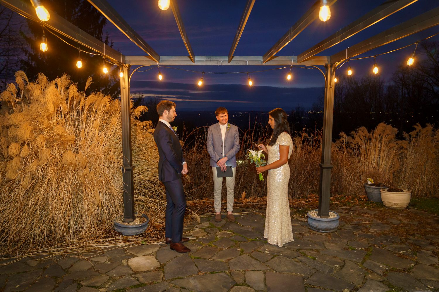 Intimate outdoor evening wedding ceremony under string lights with couple and officiant surrounded by tall ornamental grasses.