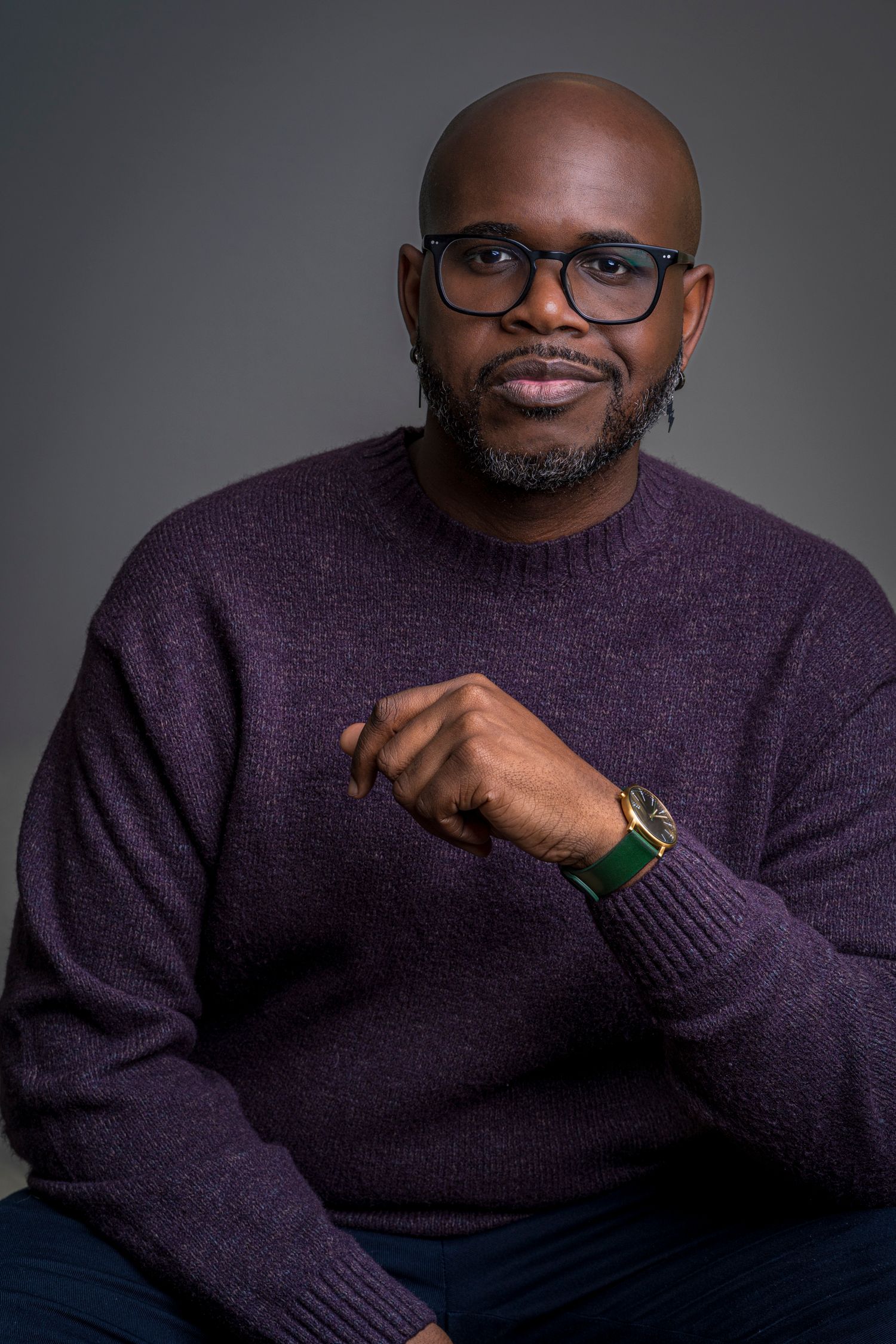 Bald Black man with glasses and beard wearing a purple sweater and watch, posing confidently against gray background.