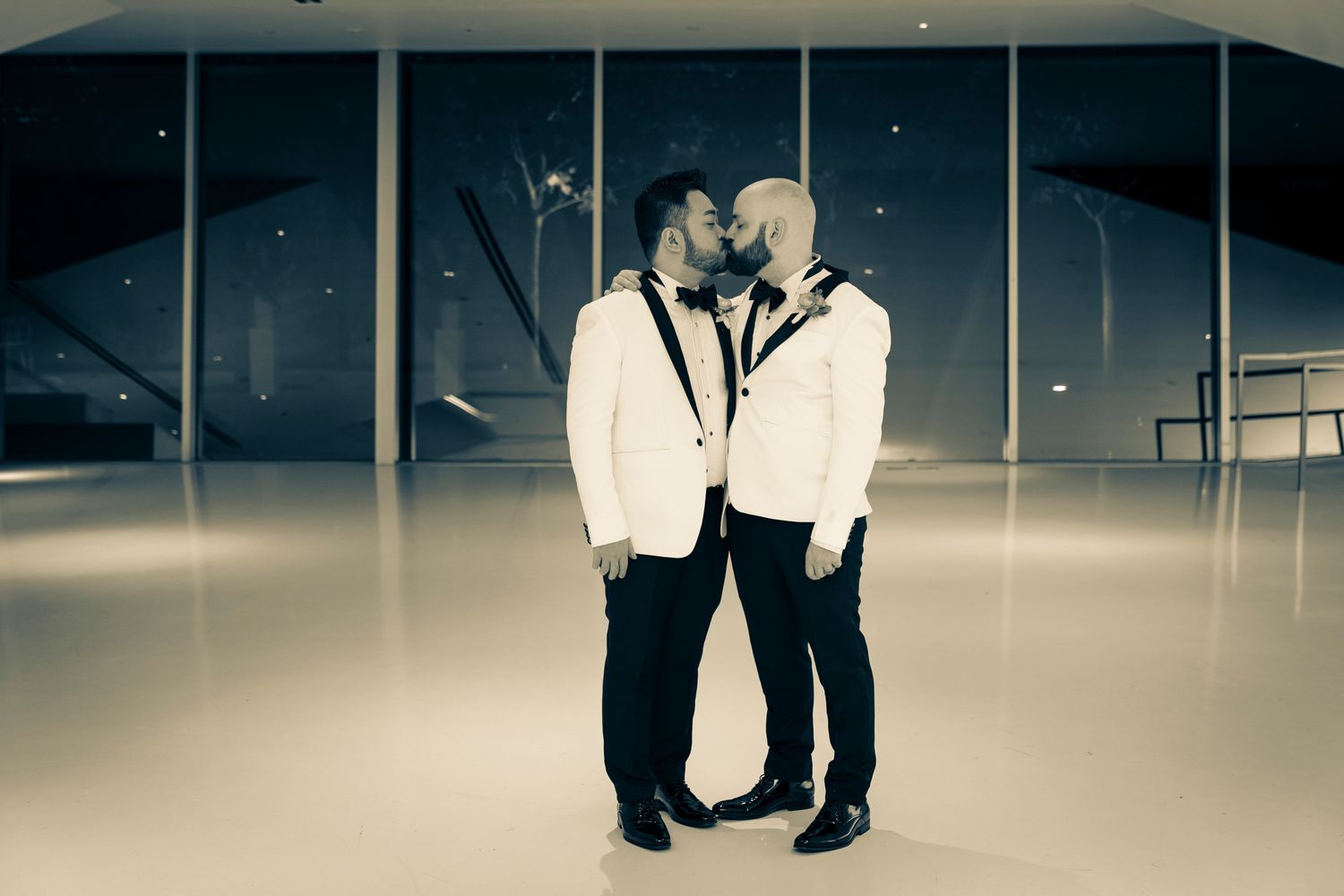 Two grooms in white tuxedos sharing romantic kiss at elegant wedding venue.